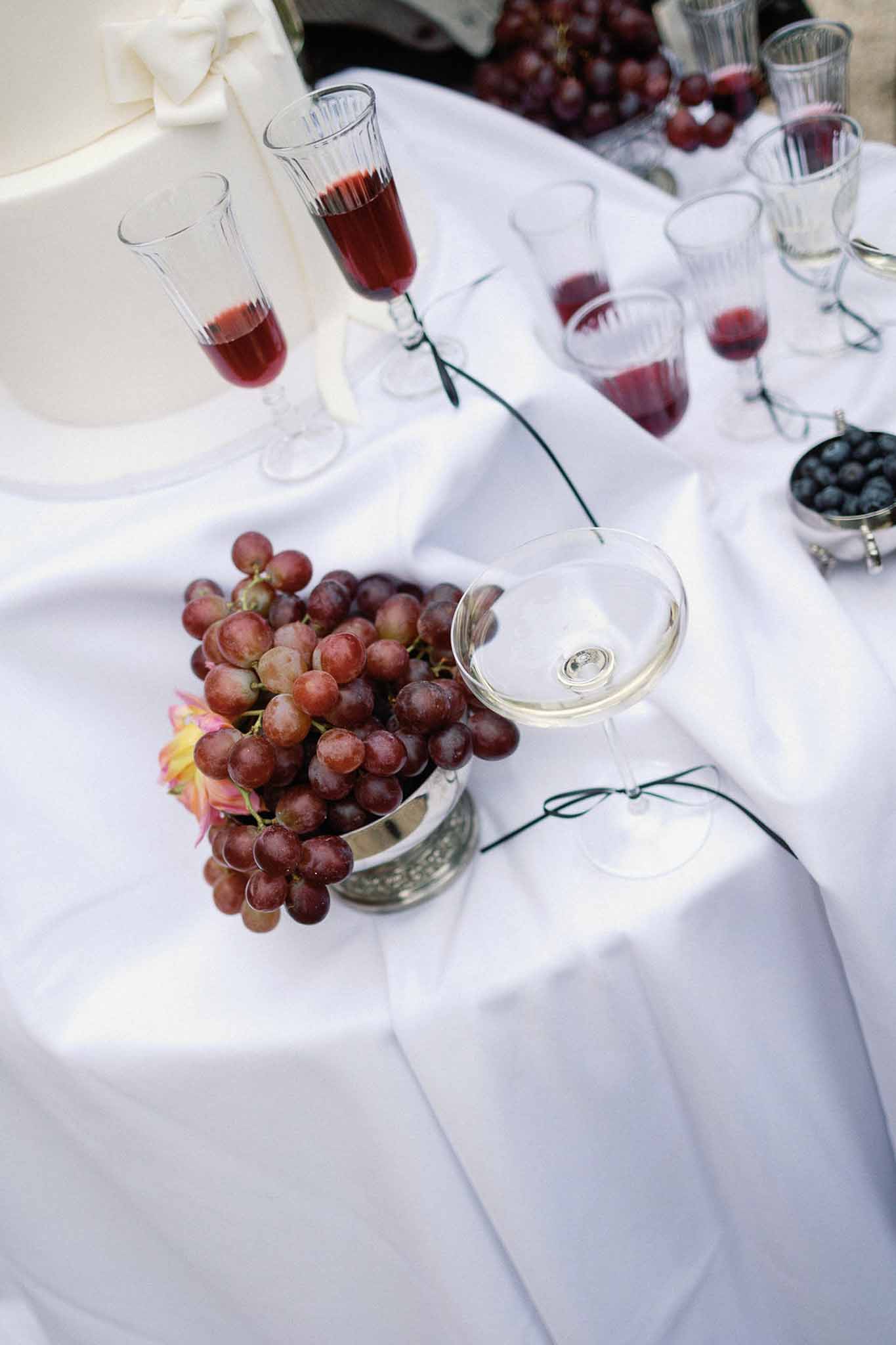 Wedding dessert table with tiered white cake, red grapes, blueberries, and champagne flutes on white linen