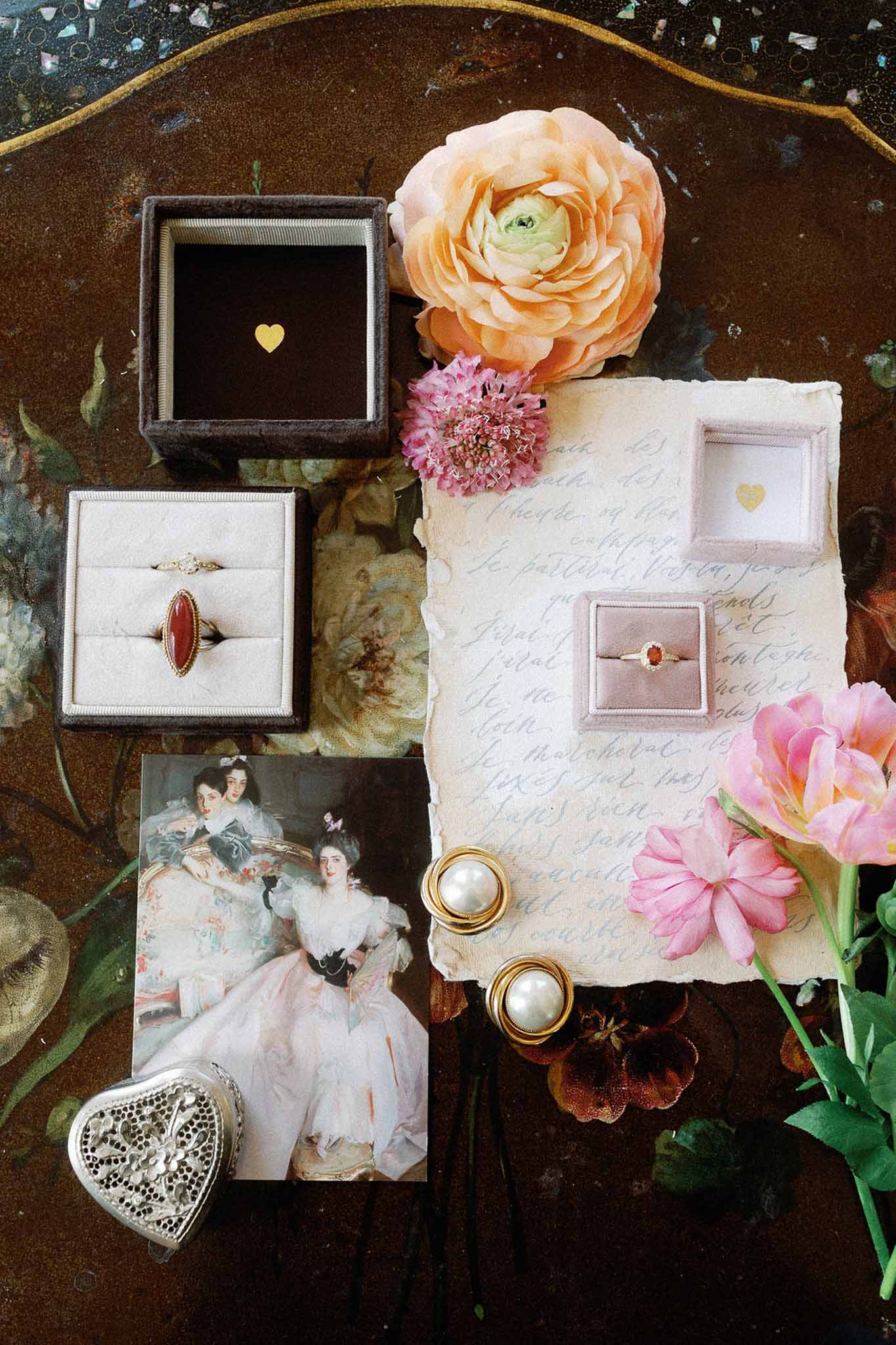 Vintage flat lay with velvet ring boxes, gold jewelry, handwritten French script, and ranunculus