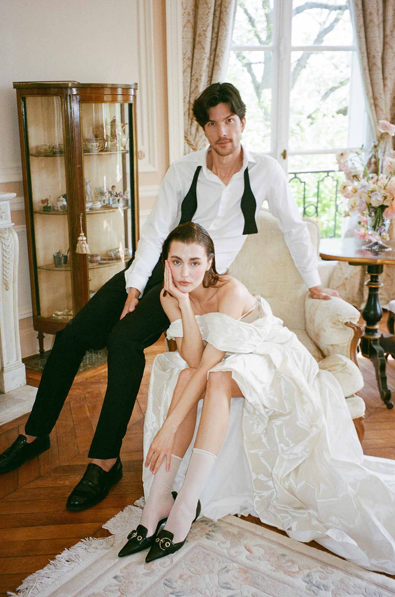 Bride in ivory ballgown seated in armchair with groom leaning behind in wood-panelled chateau salon