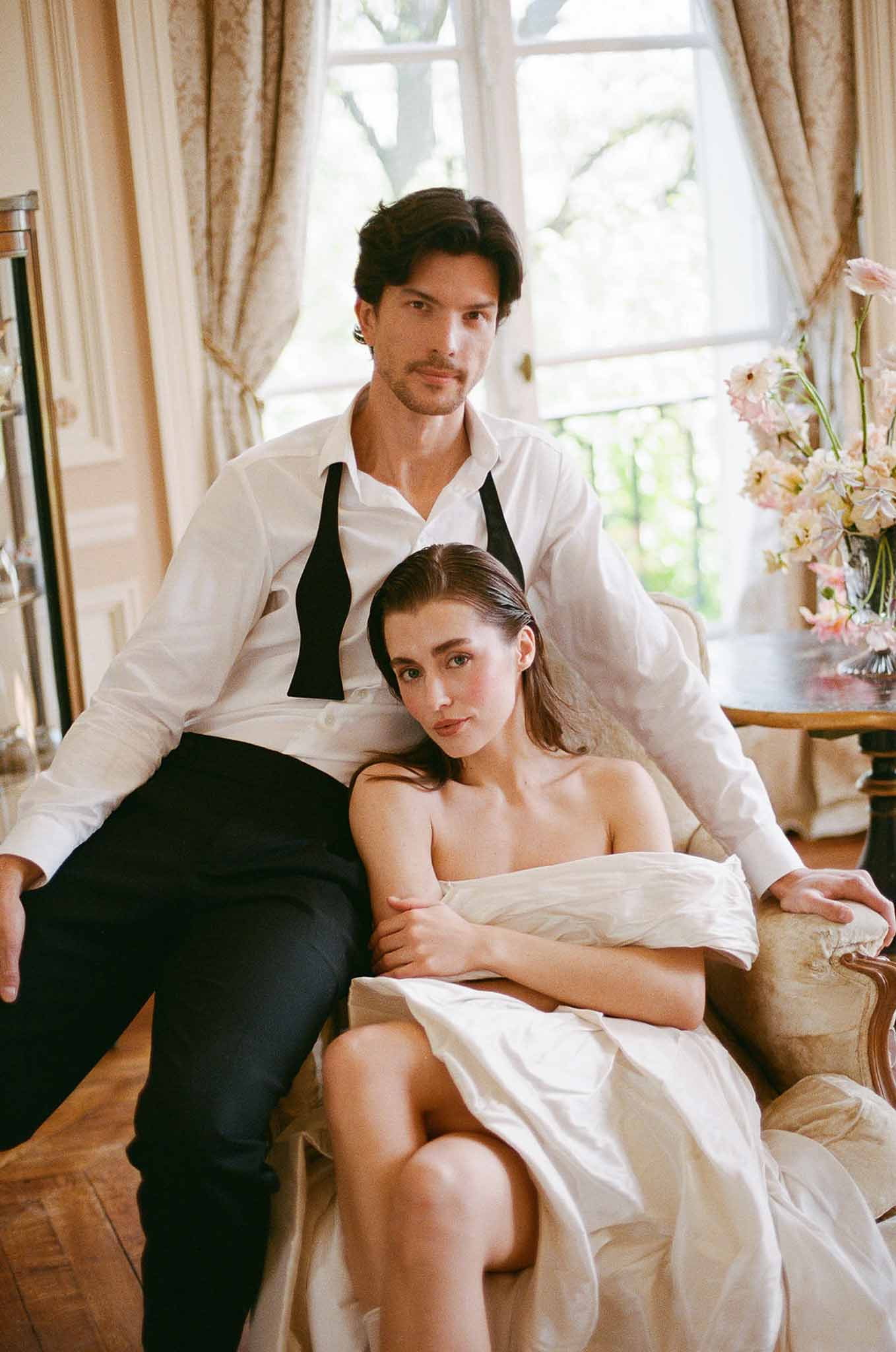 Groom standing behind seated bride in gilt armchair in boiserie salon with draped ivory curtains