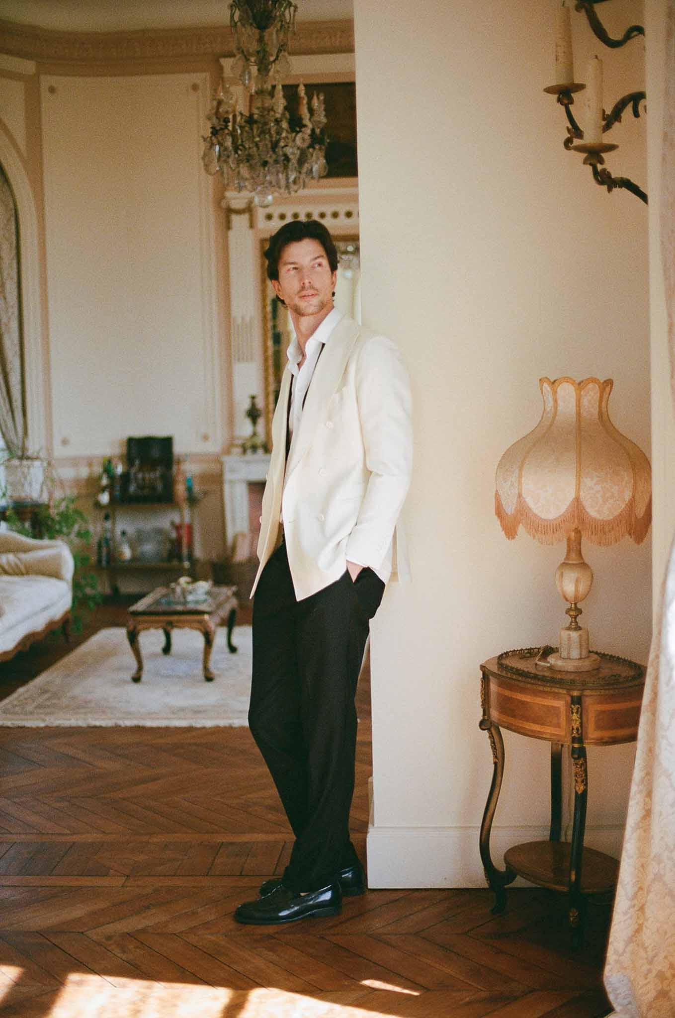 Groom in ivory blazer and black trousers leaning against column in Parisian salon with chandelier and gilt fireplace