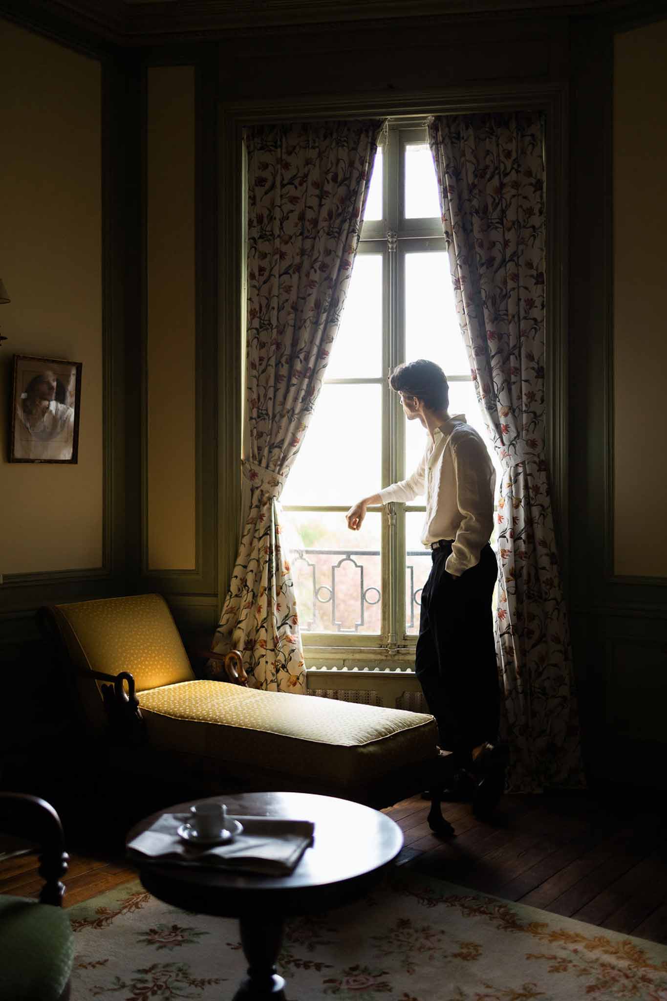 Groom in white shirt looks out window in olive-paneled room with mustard chaise and floral curtains