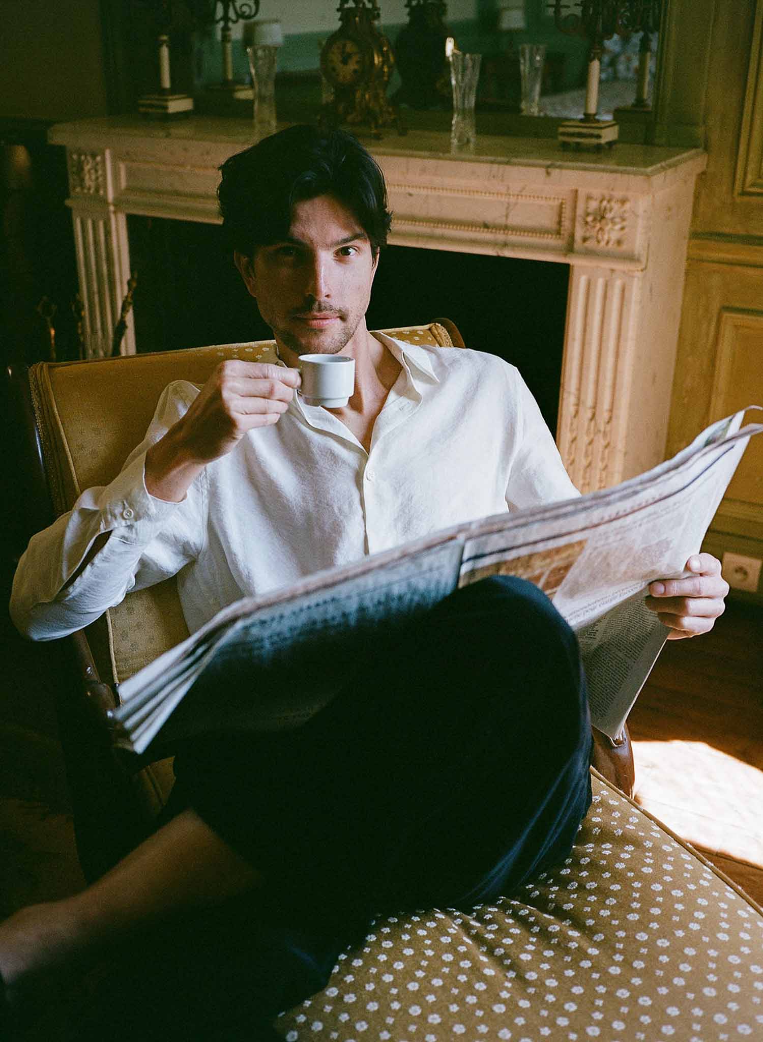 Groom in white linen shirt reading newspaper with espresso in armchair beside chateau stone fireplace