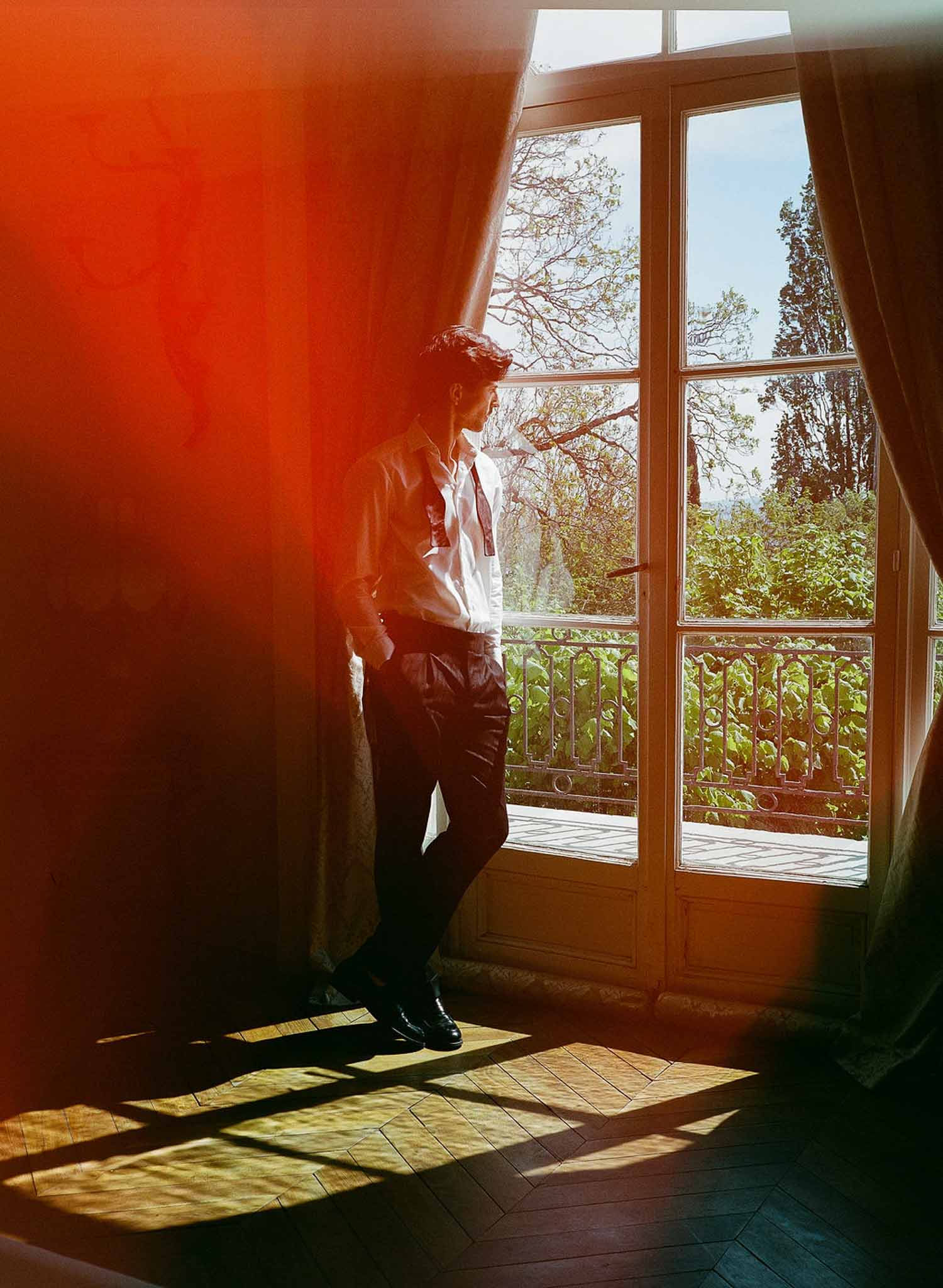 Groom in white shirt and dark trousers by open French doors with dramatic backlight and warm lens flare