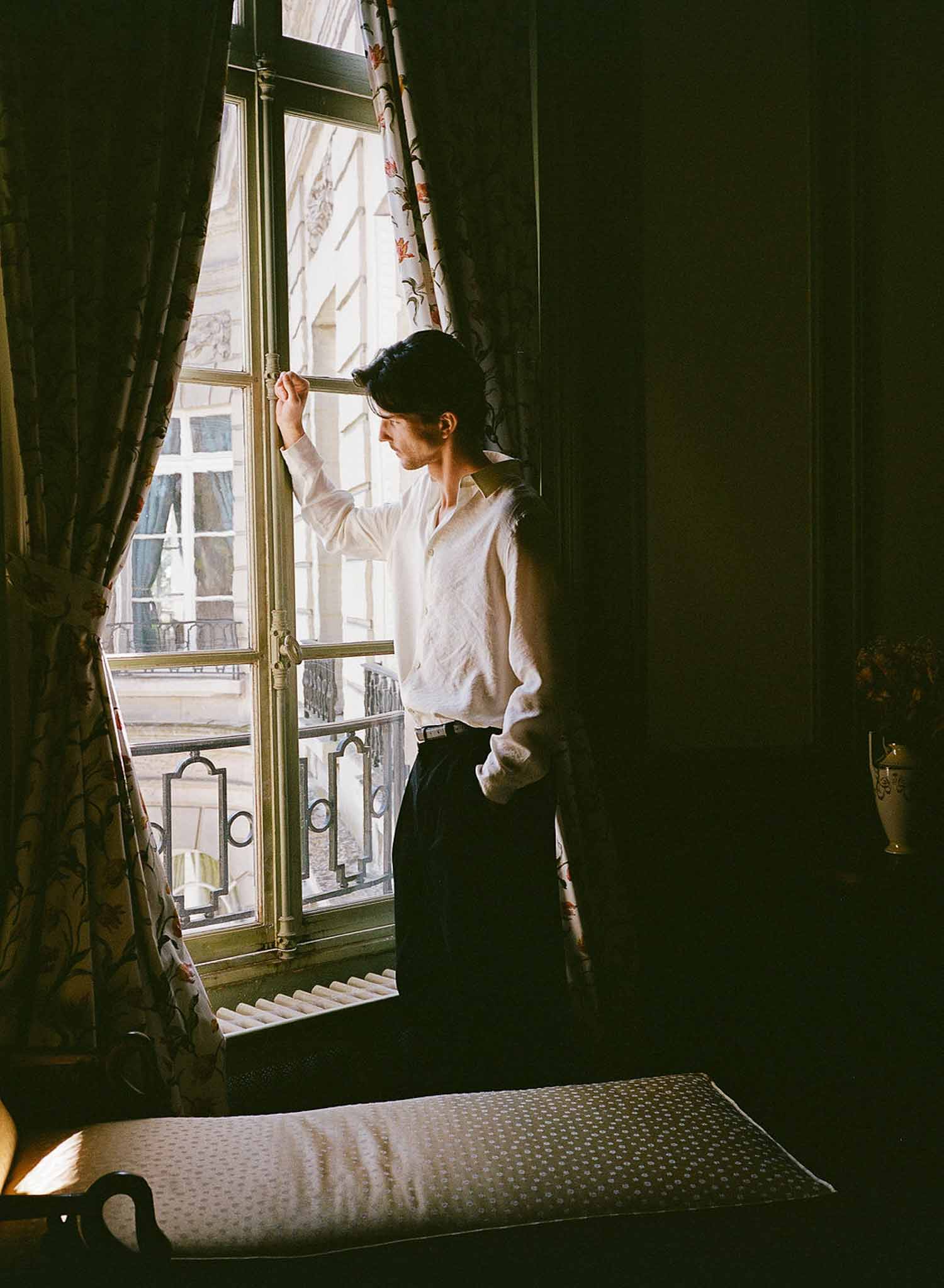 Groom in cream linen shirt at tall French window with gilded frame in Parisian hotel room