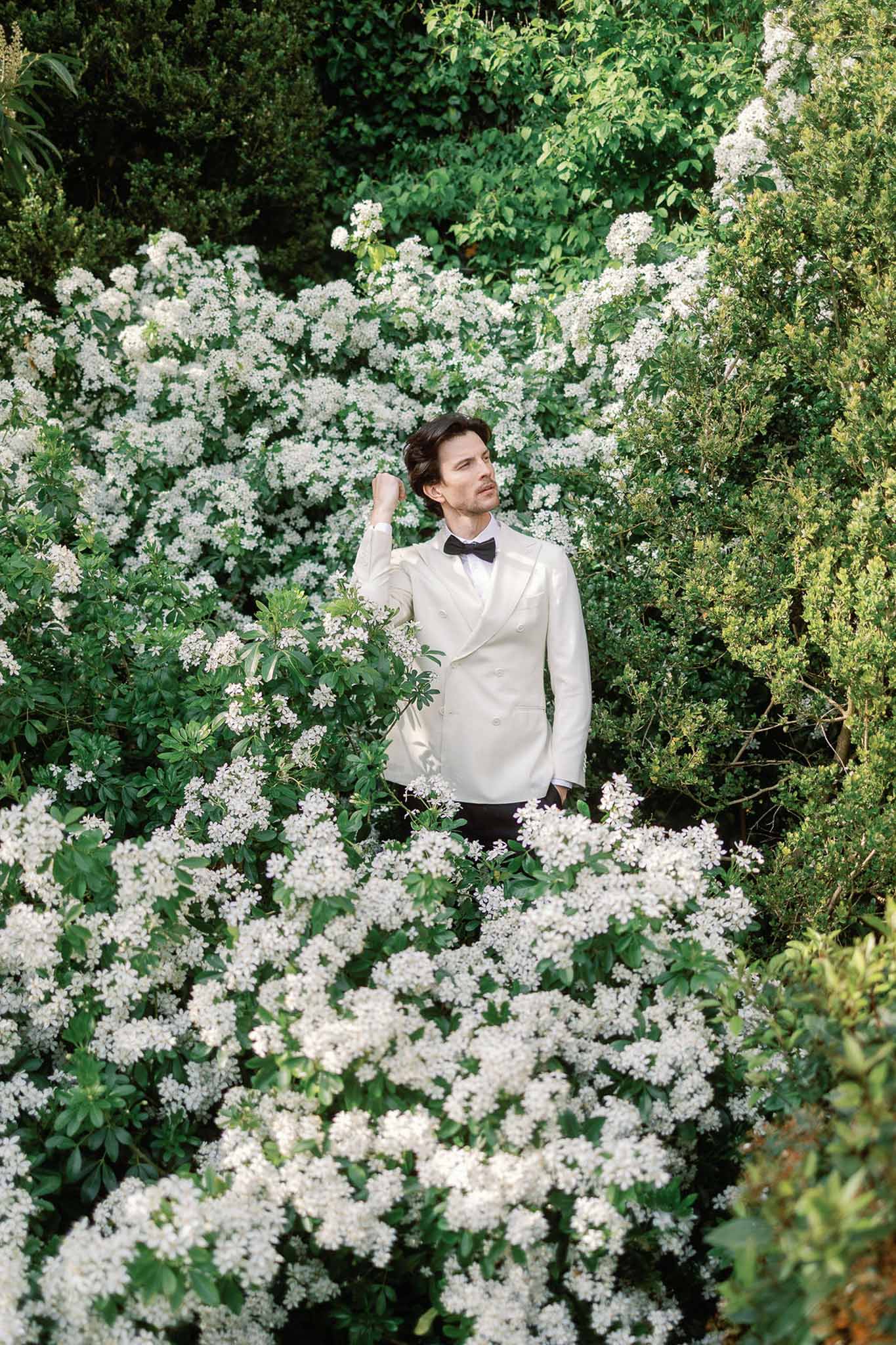 Groom in ivory double-breasted dinner jacket and black bow tie standing among white flowering shrubs in garden