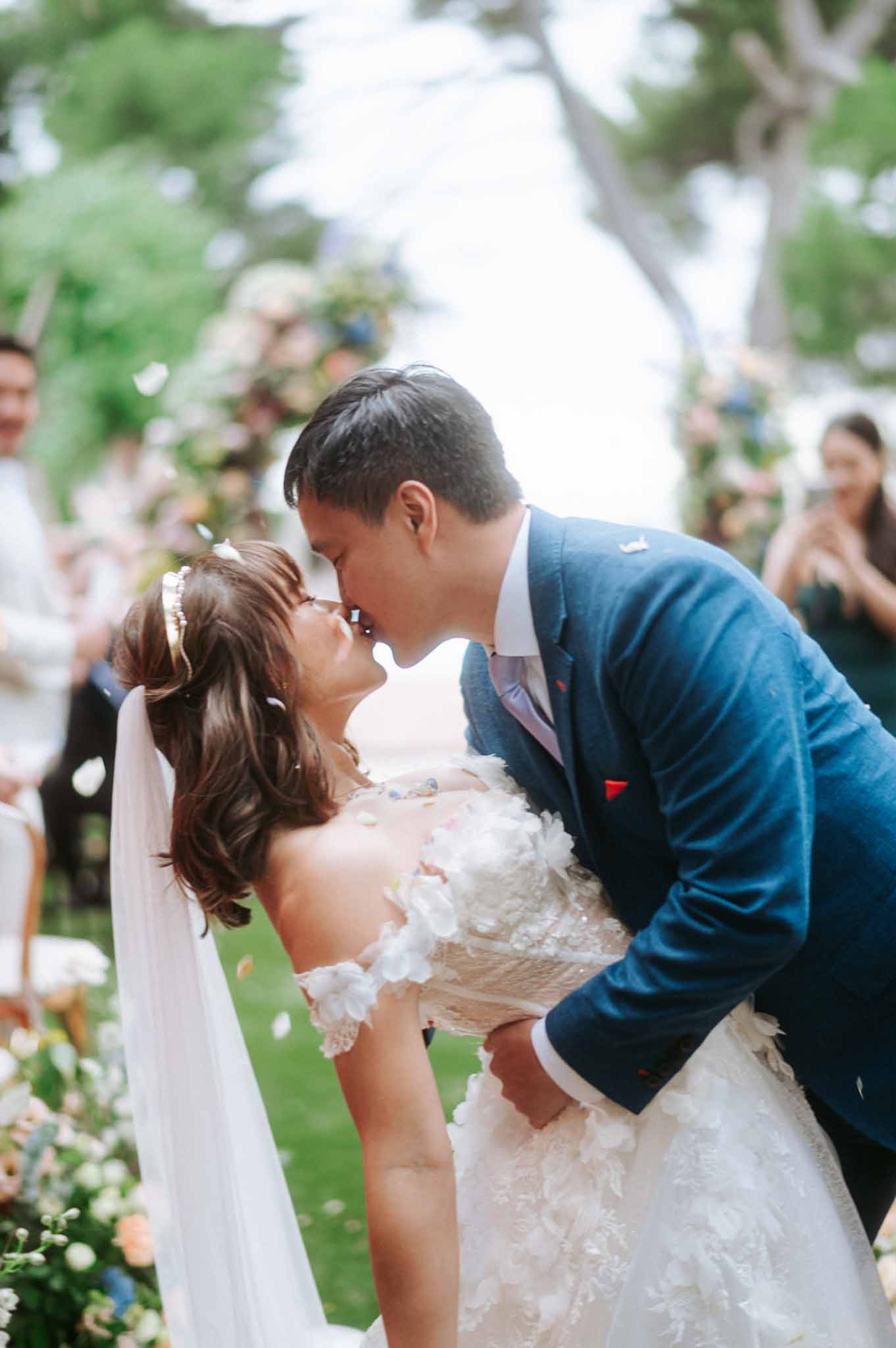 Groom dipping bride for first kiss as confetti falls at garden ceremony with floral arch in background