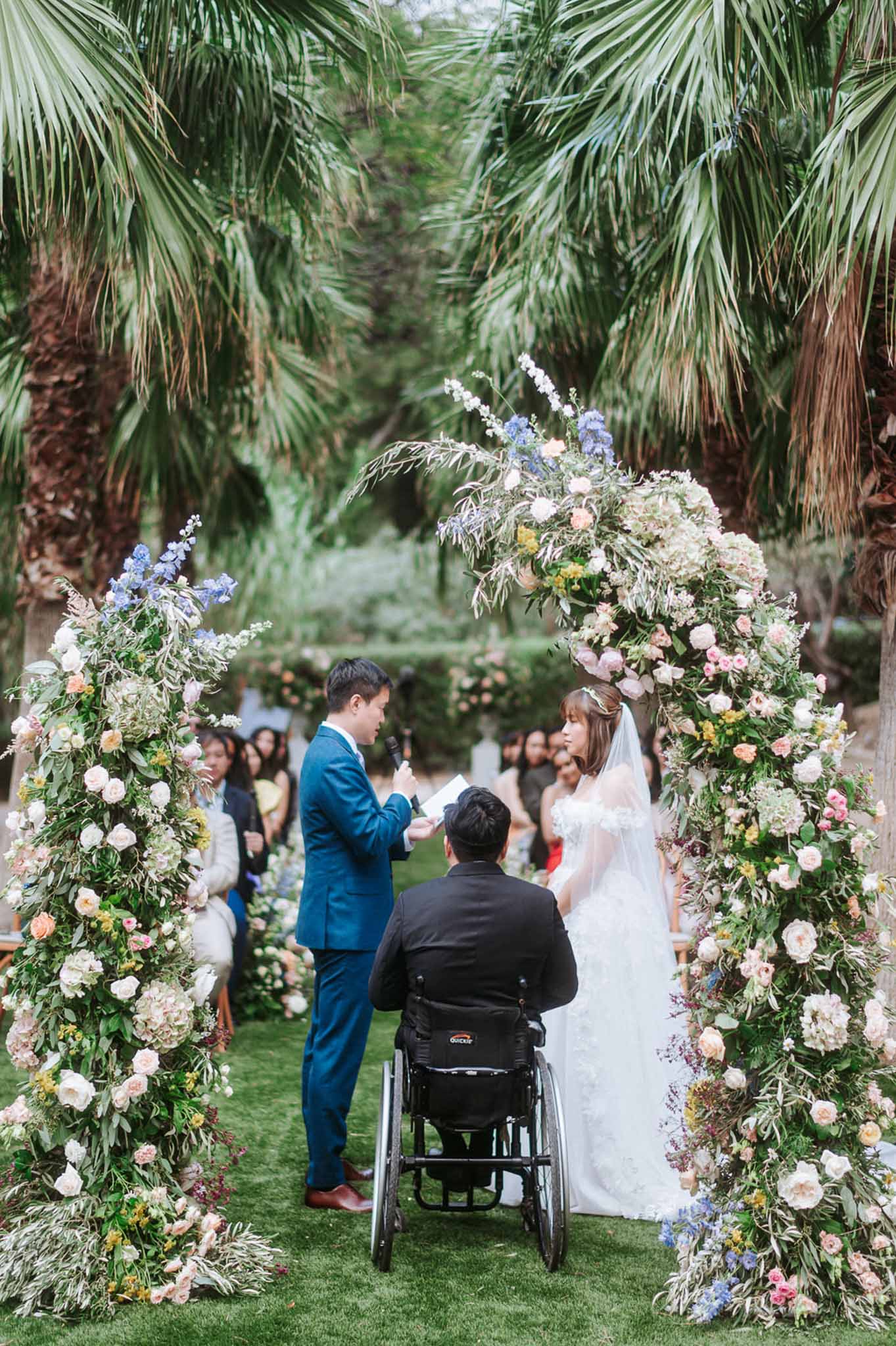 Outdoor ceremony under a circular floral arch of blush roses and blue delphiniums with palm trees and seated guests