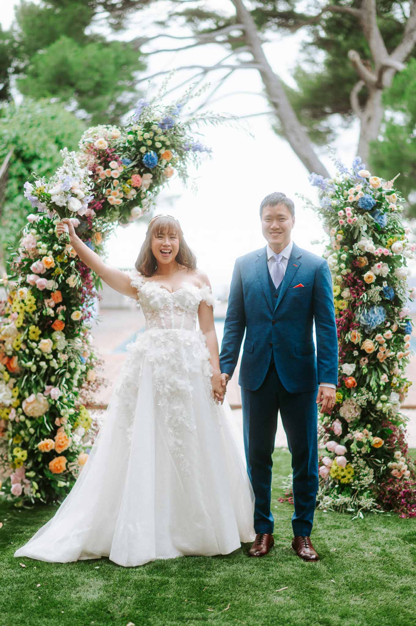 Bride and groom holding hands before circular floral arch of peach roses and blue hydrangeas on outdoor lawn