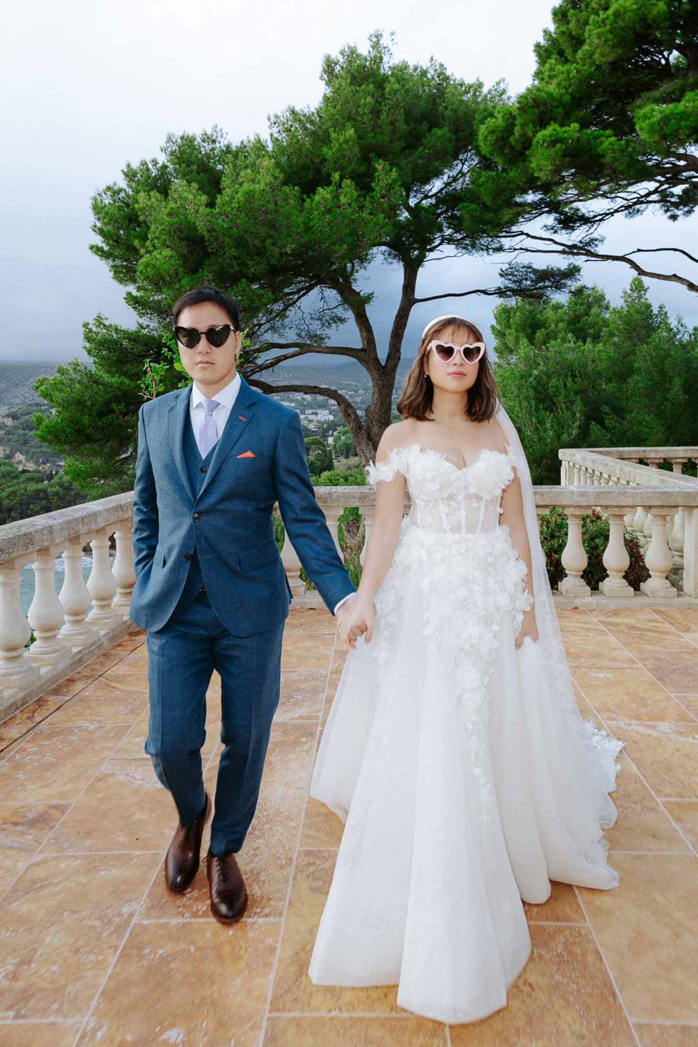 Bride and groom walking on coastal terrace wearing heart-shaped sunglasses, editorial playful portrait