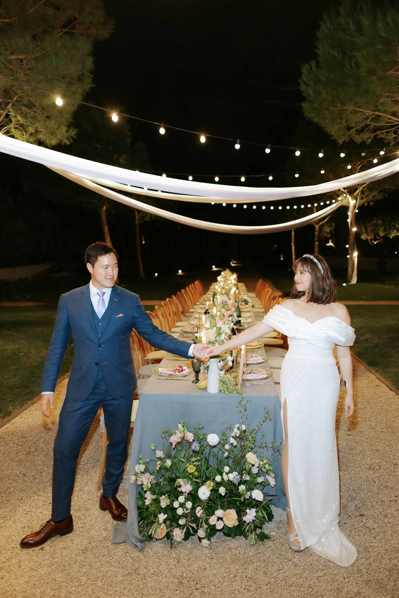 Couple at head of long table with peach garland centrepiece and globe lights on gravel at night