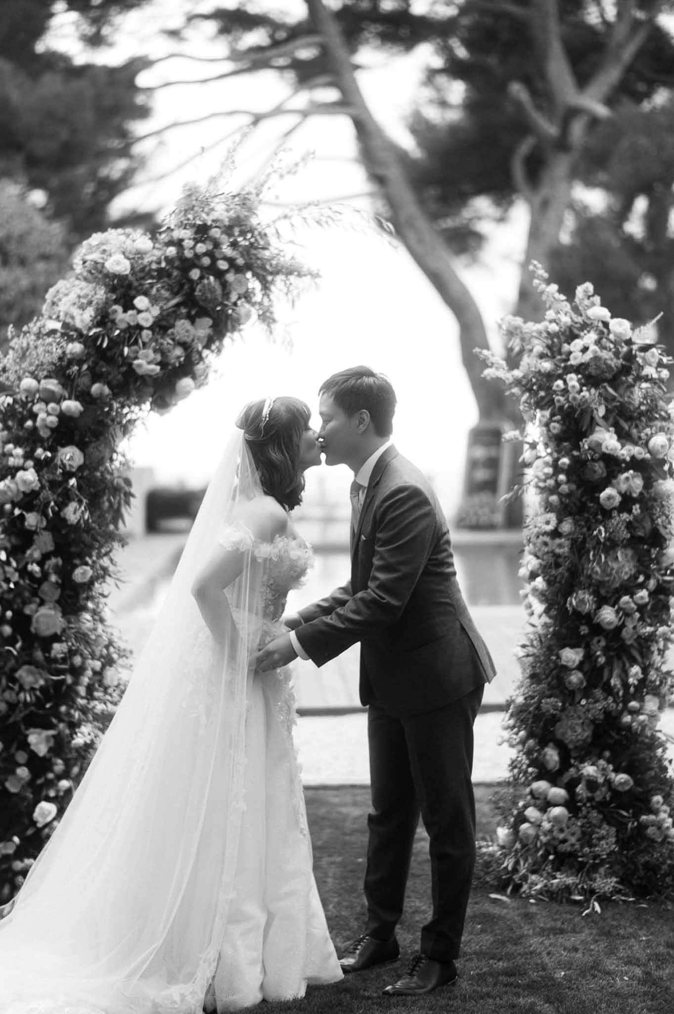 Black and white first kiss under dense circular floral arch in garden ceremony