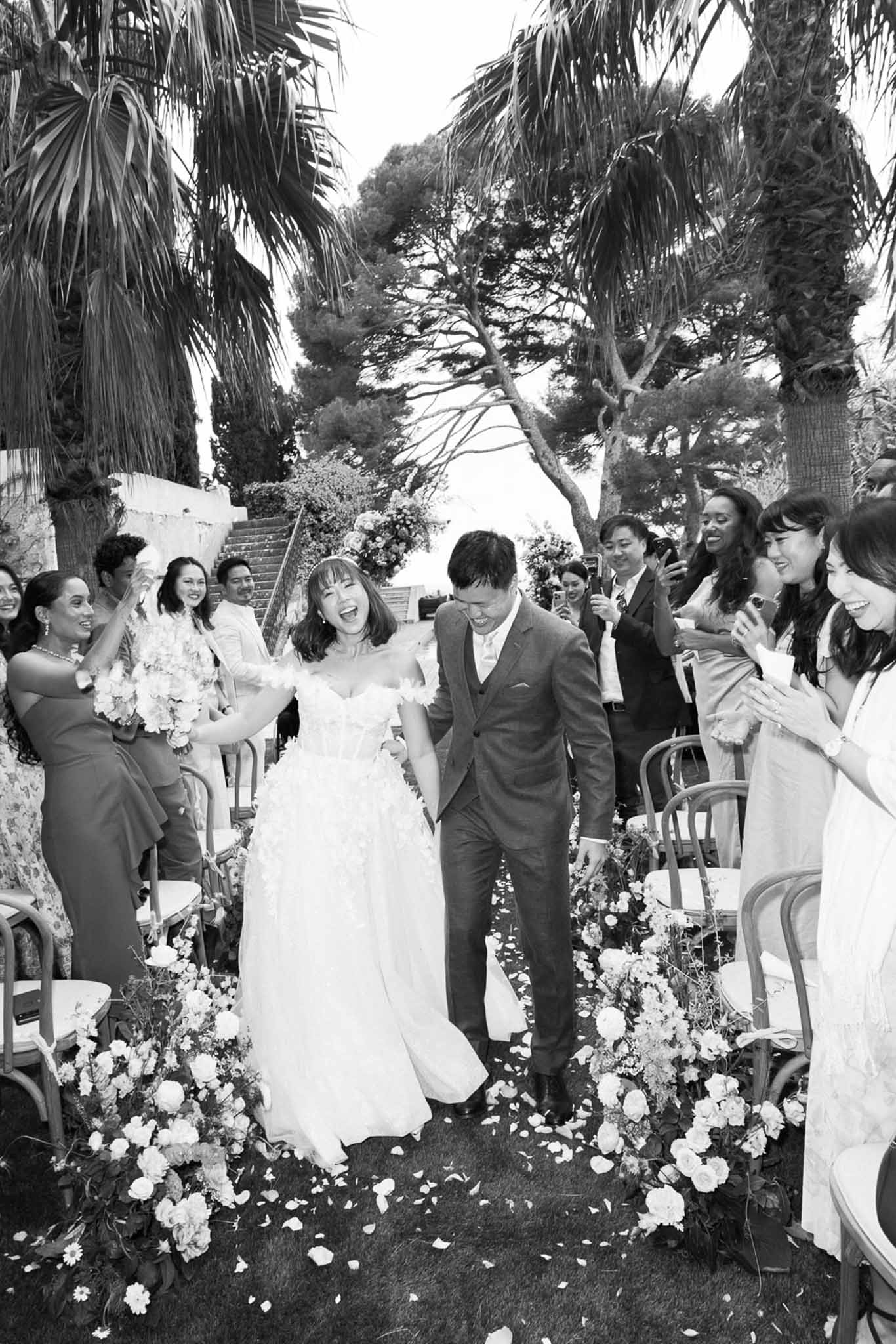 Black and white photo of bride and groom walking back down the aisle as guests clap and cheer at outdoor garden ceremony