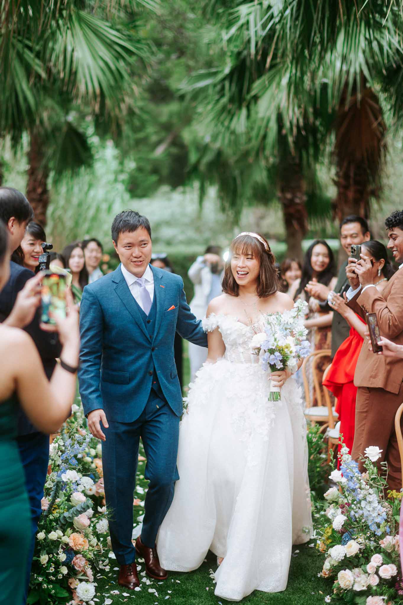 Couple walking back down palm-lined aisle past blue delphinium and blush rose arrangements