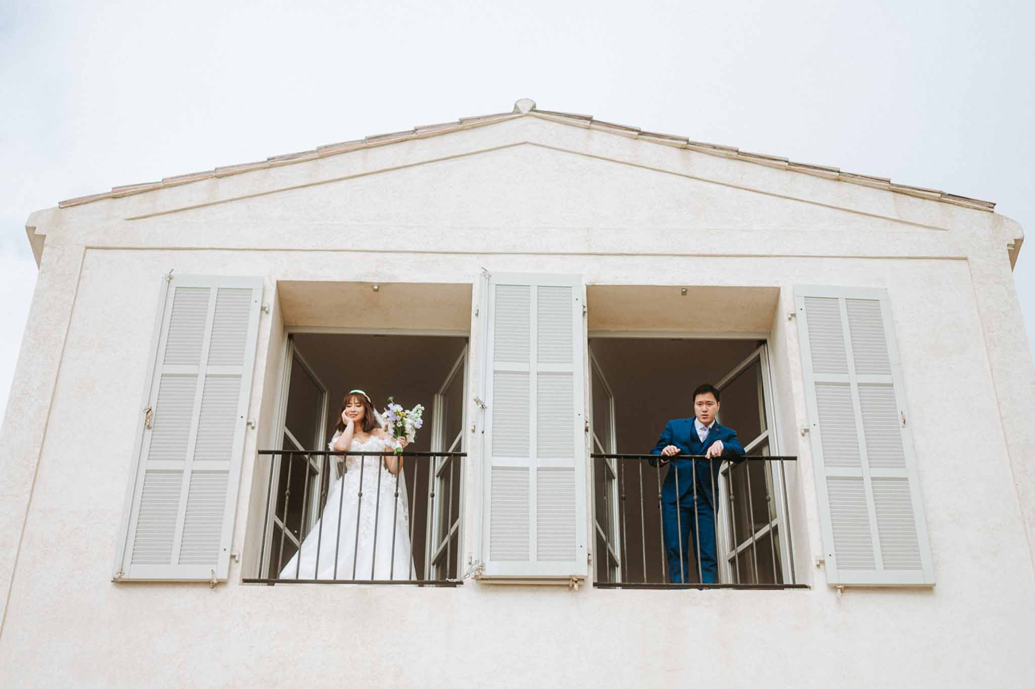 Bride and groom on adjacent balconies of cream Provencal building with louvred shutters and pediment
