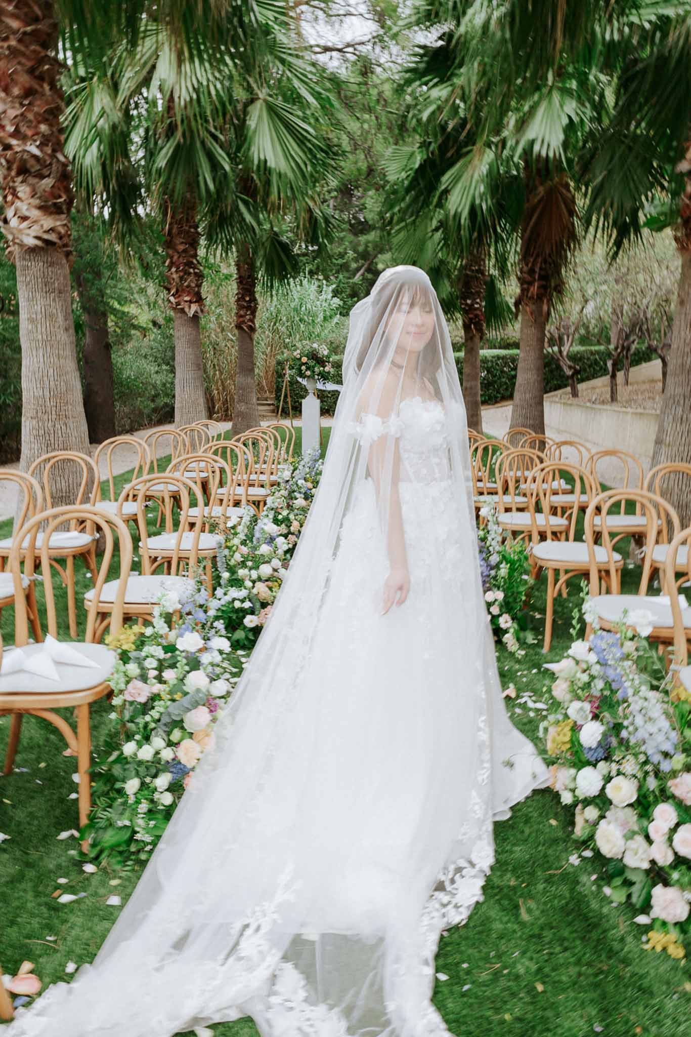 Bride in applique ballgown with cathedral veil on palm-tree garden aisle lined with pastel floral installation