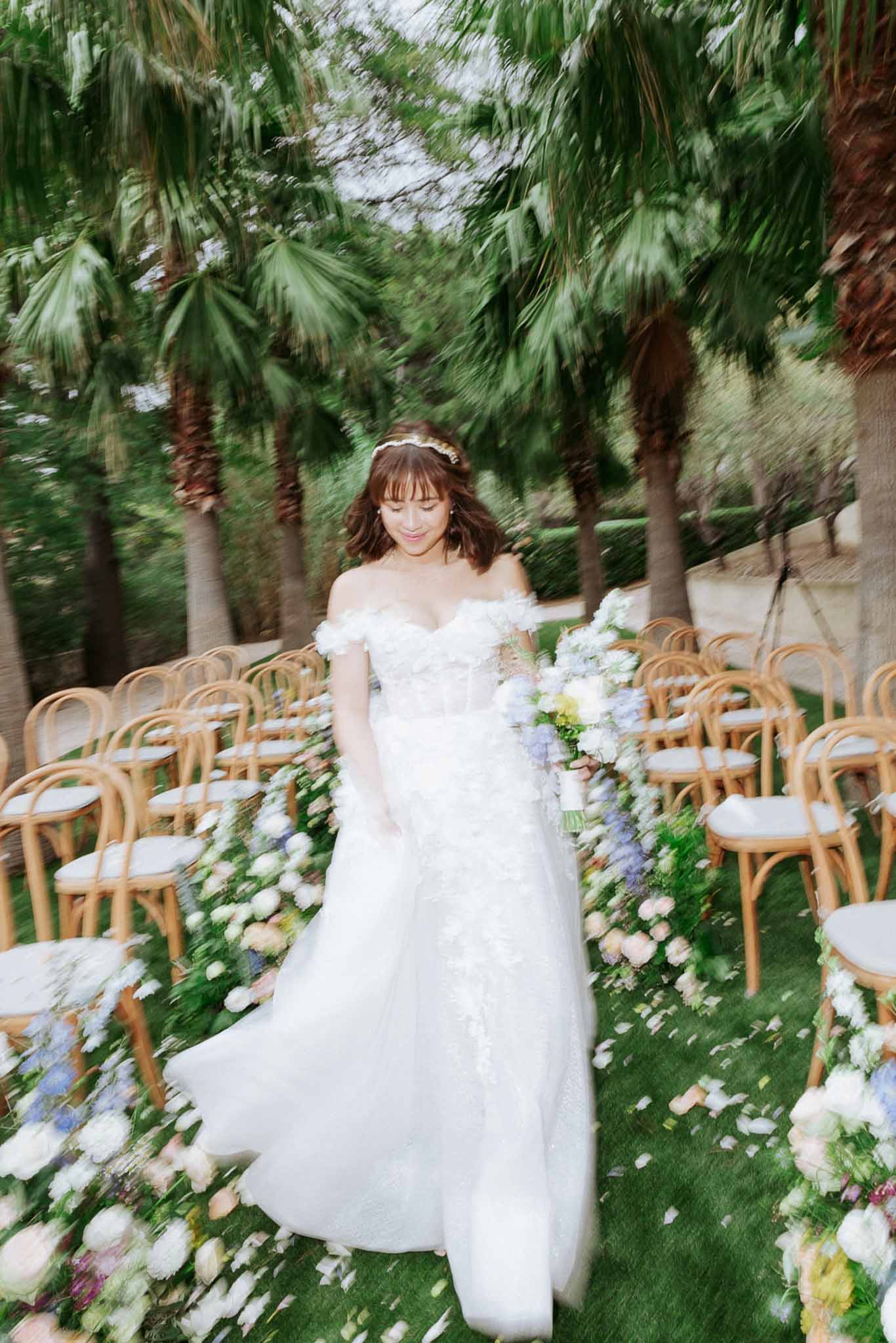 Bride in off-shoulder gown walks aisle with motion blur past white, blush, and blue floral arrangements