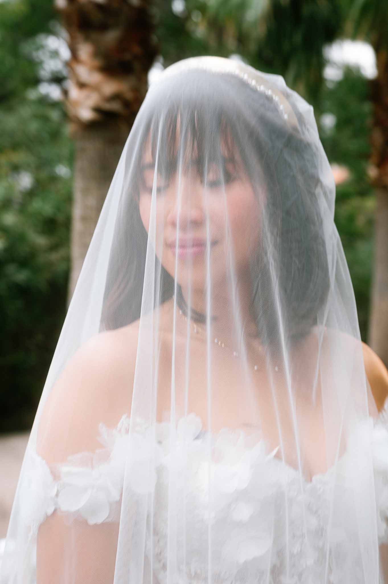 Close-up bride smiling through sheer tulle veil wearing strapless floral applique gown and pearl necklace