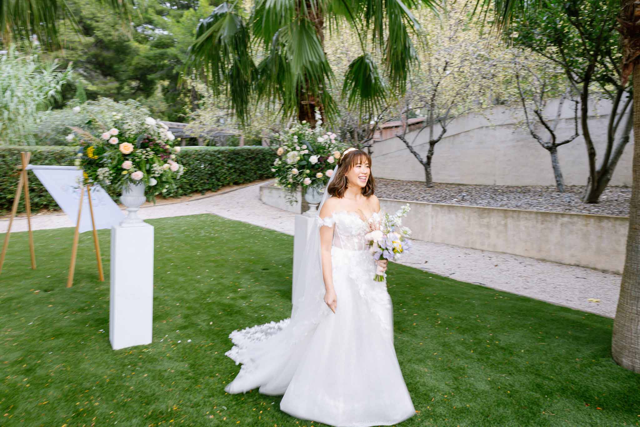 Bride walking with lavender bouquet past white pedestal urns of blush and peach flowers on garden lawn