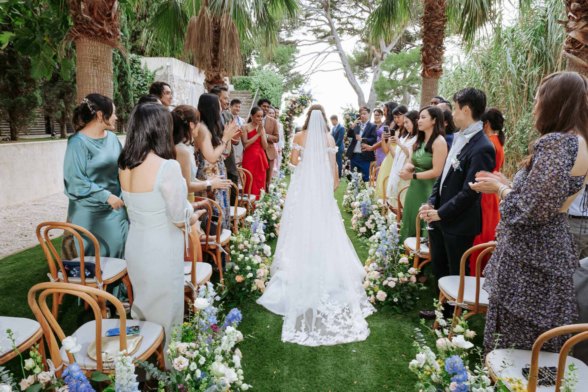 Bride walking down an outdoor aisle with lace-hemmed cathedral veil as guests stand and clap beside floral arrangements