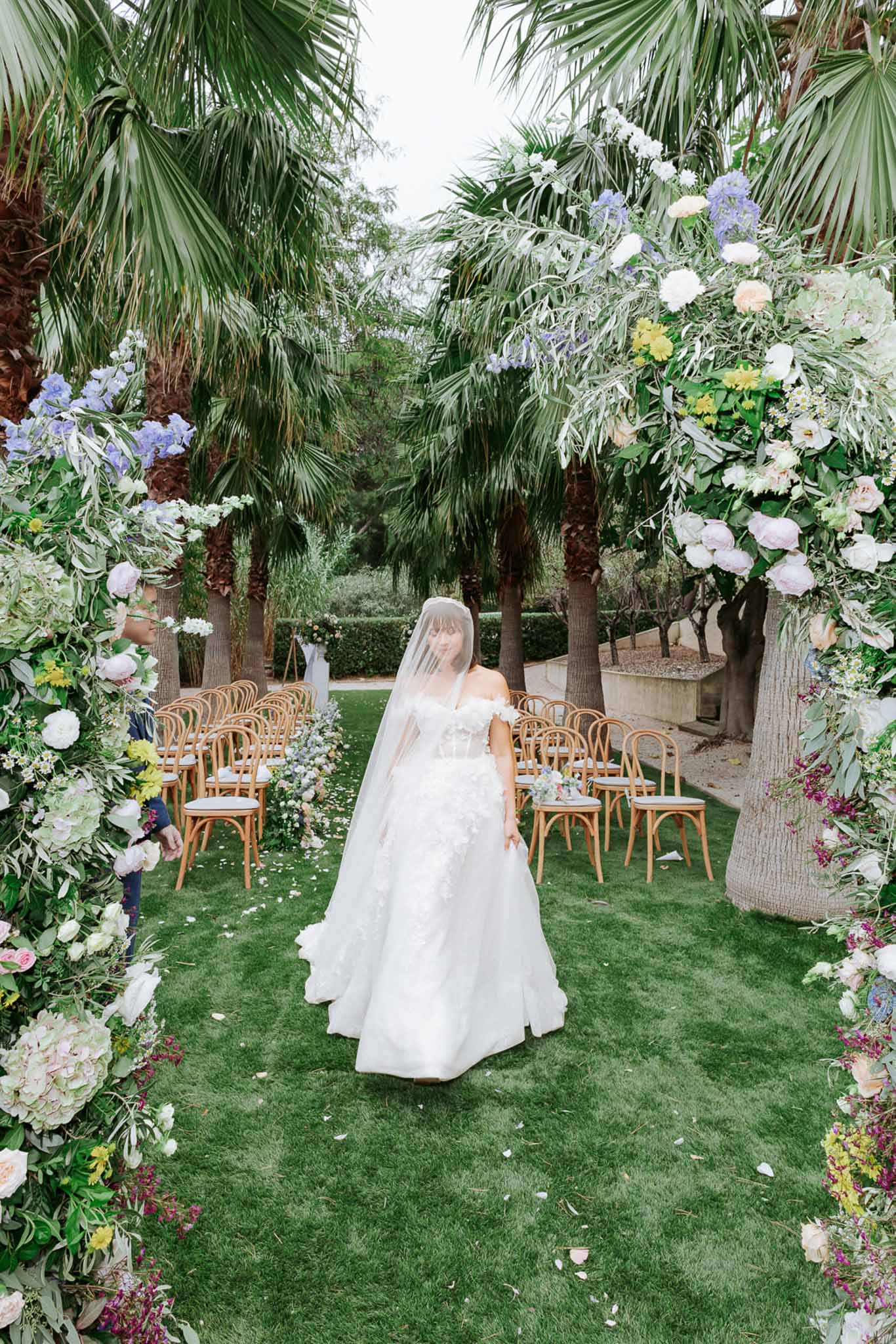 Bride walks down outdoor aisle through floral arch of roses, peonies, and hydrangeas on palm-lined lawn