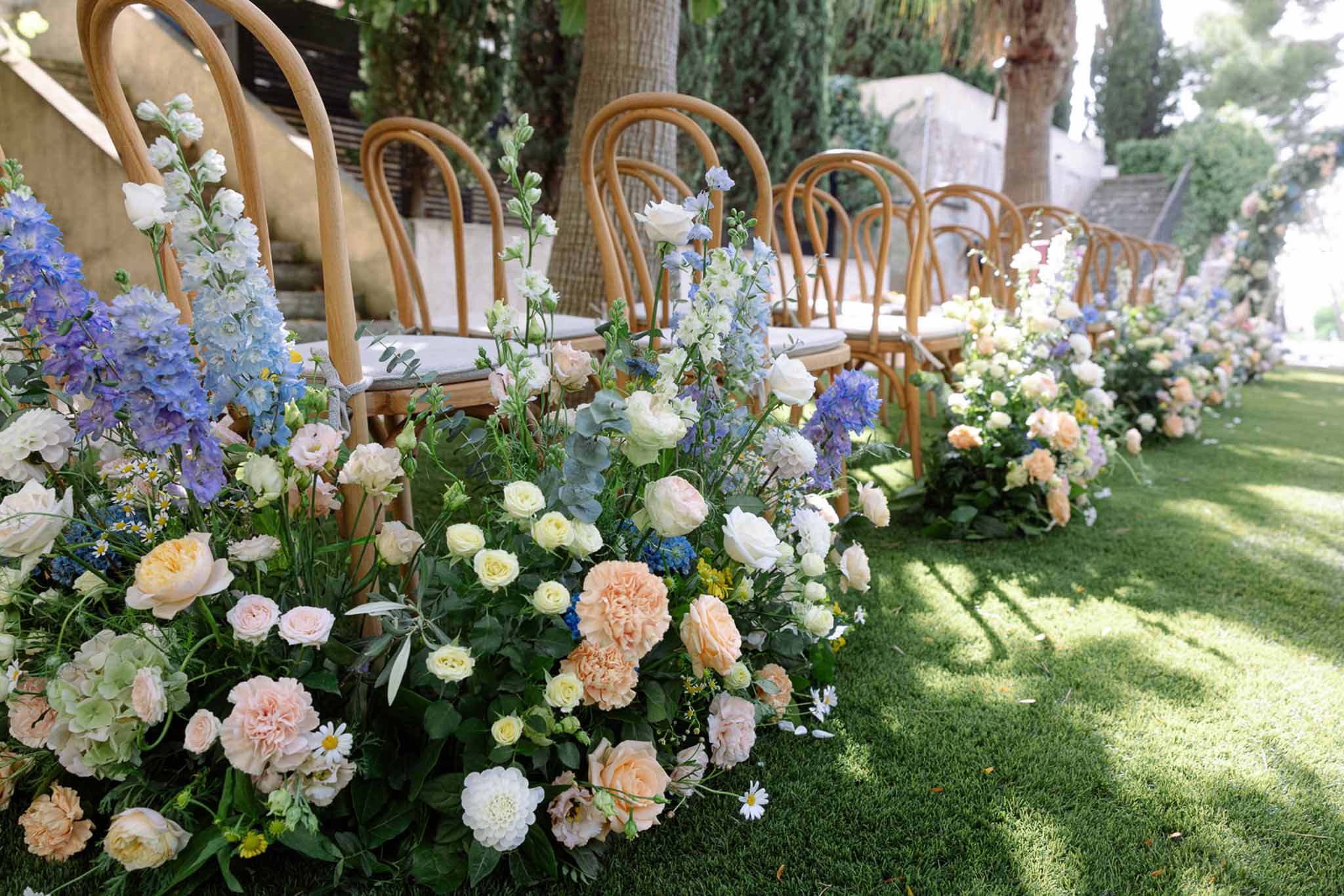 Ceremony aisle lined with rattan chairs and pastel floral arrangements of blush roses and blue delphiniums on lawn