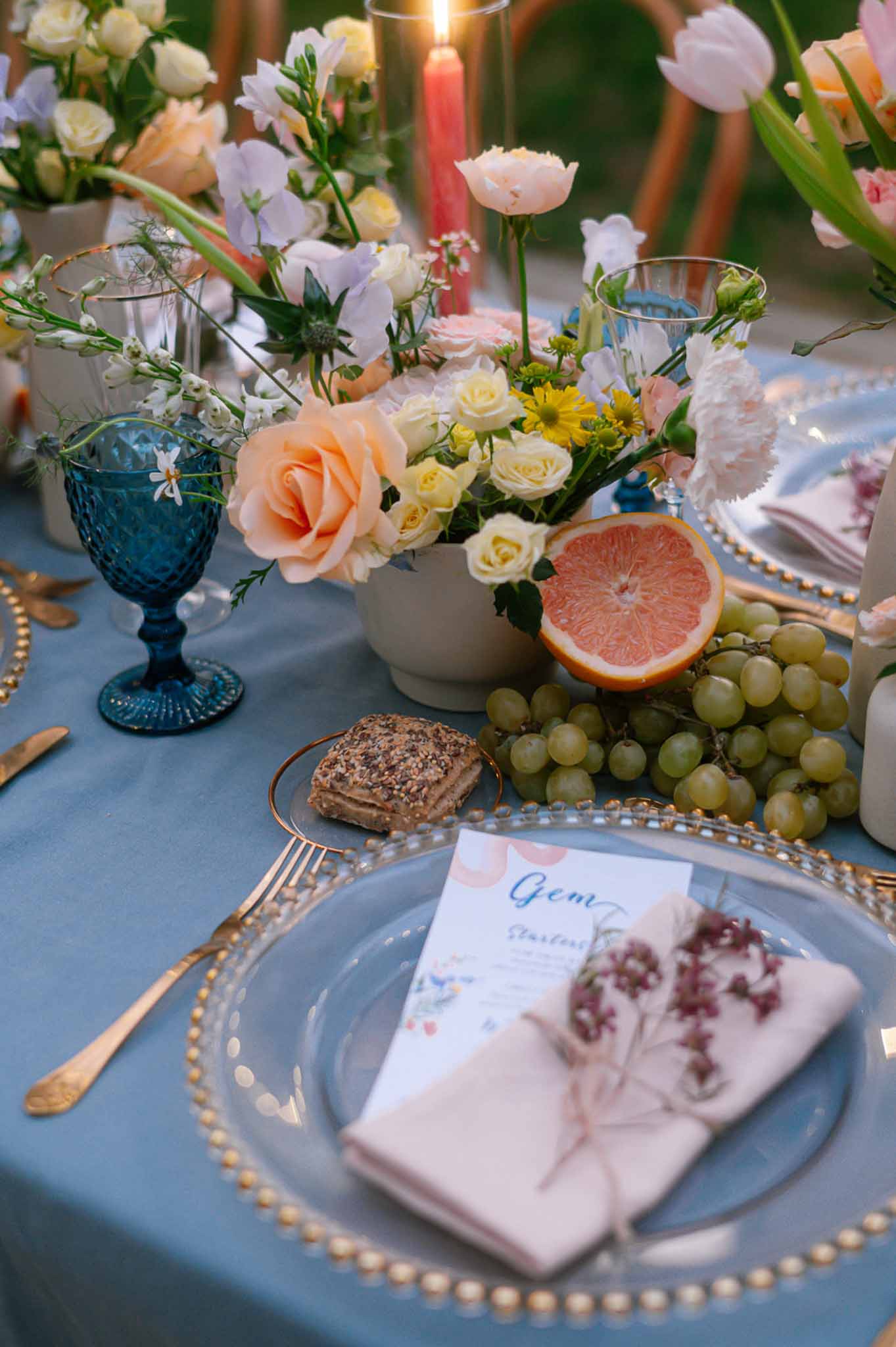 Colorful reception tablescape with dusty blue linen, gold flatware, peach roses, and coral taper candles