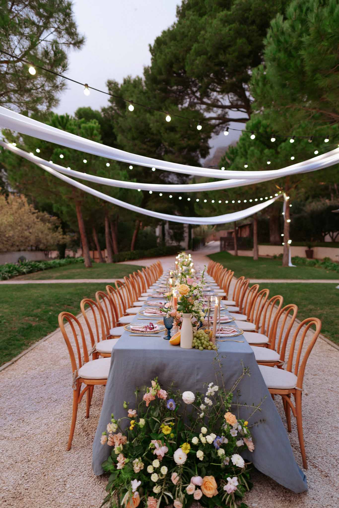 Long feasting table with dusty blue linen, peach and blush florals, and globe lights under tree canopy