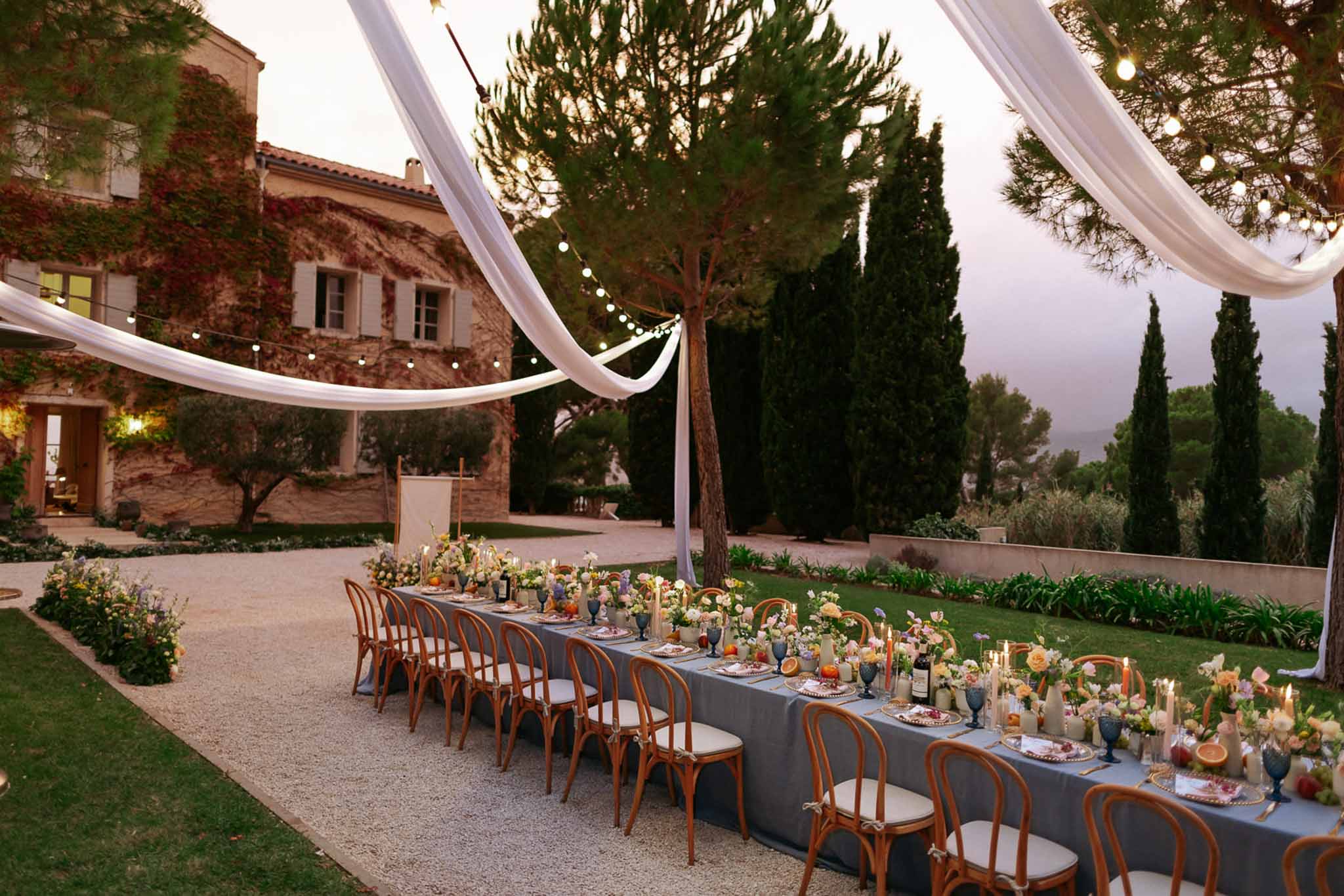 Dusk reception table with slate-blue linen and mixed floral arrangements under draped canopy at a Provencal mas