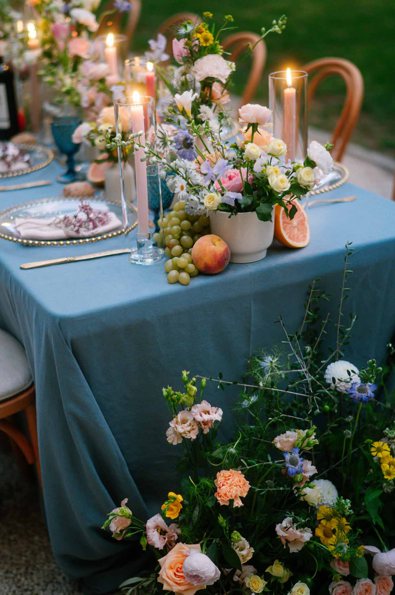 Dusty blue linen table with garden rose centrepiece, fresh fruit, gold flatware, and cobalt glassware