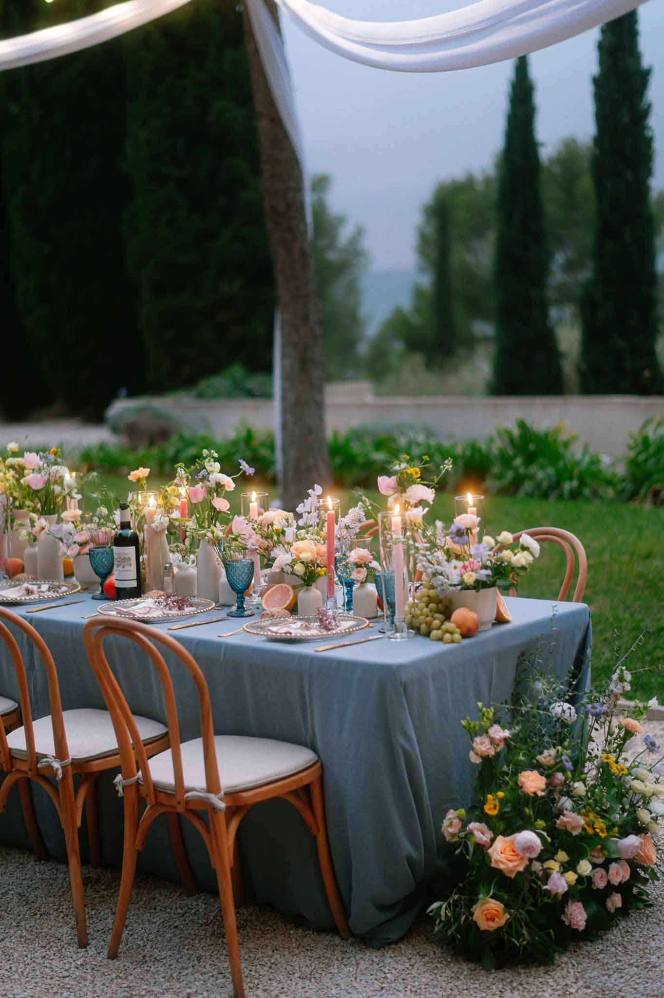 Long reception table with dusty blue linen, coral taper candles, and peach and blush roses in ceramic bud vases at dusk