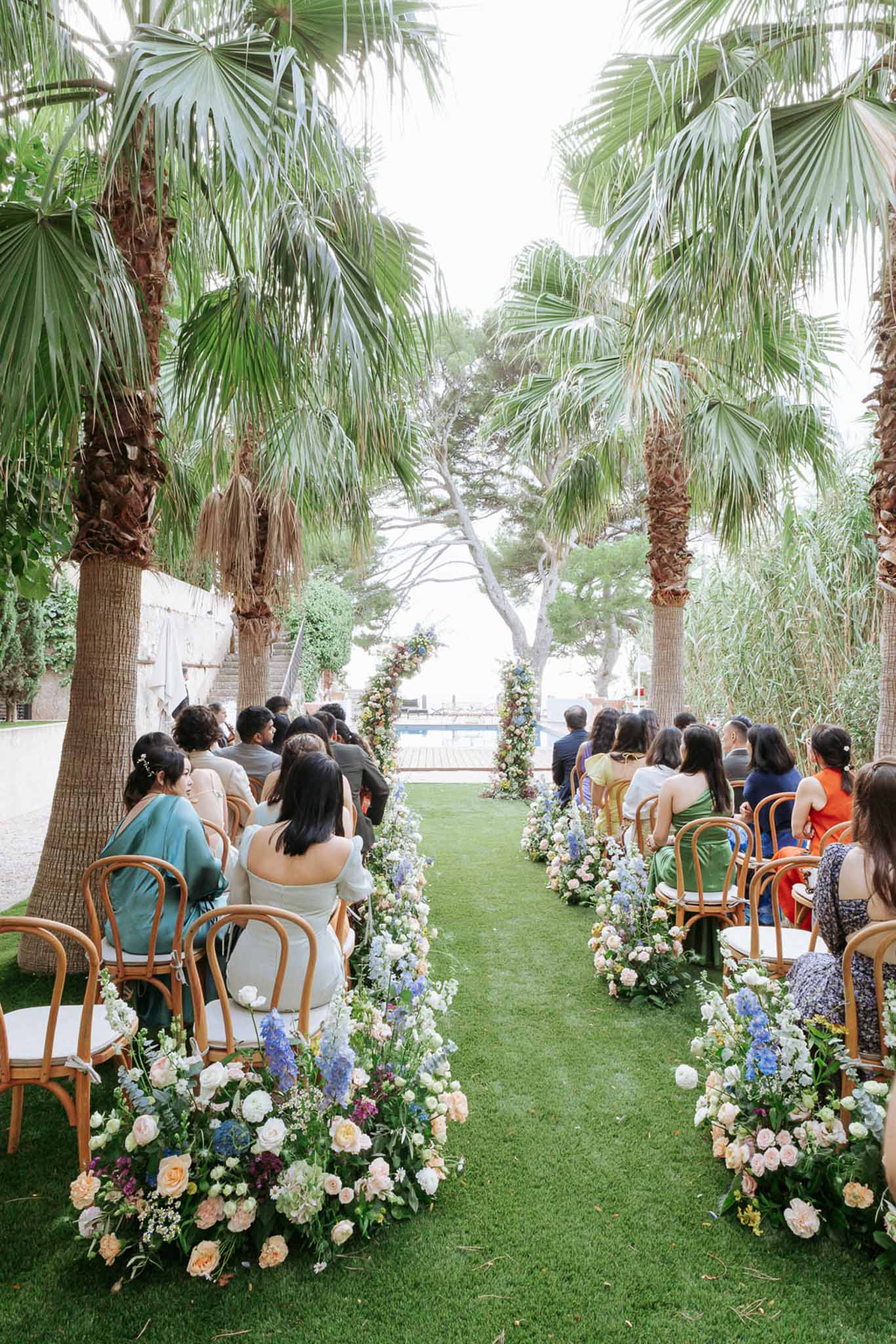 Garden wedding ceremony with guests seated on rattan chairs, peach and blue floral aisle arrangements, and palm-framed altar