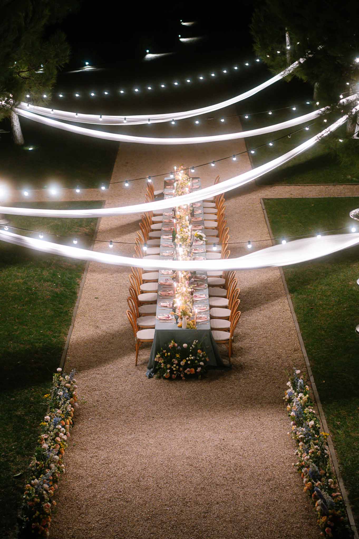 Aerial view of long sage-green table with peach floral runner, draped canopy, and globe string lights
