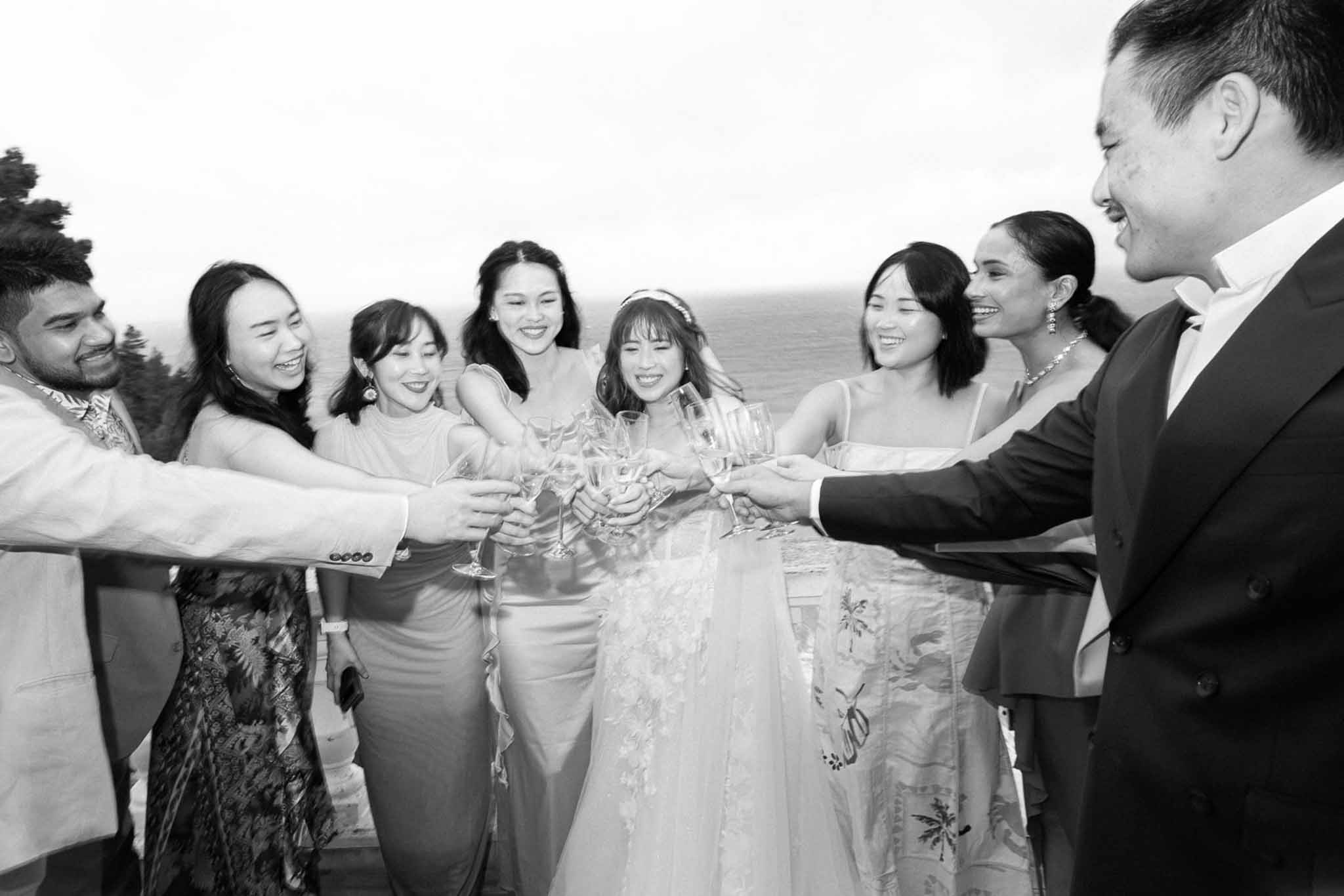 Bride and eight friends clink champagne flutes on elevated terrace with water view in B&W
