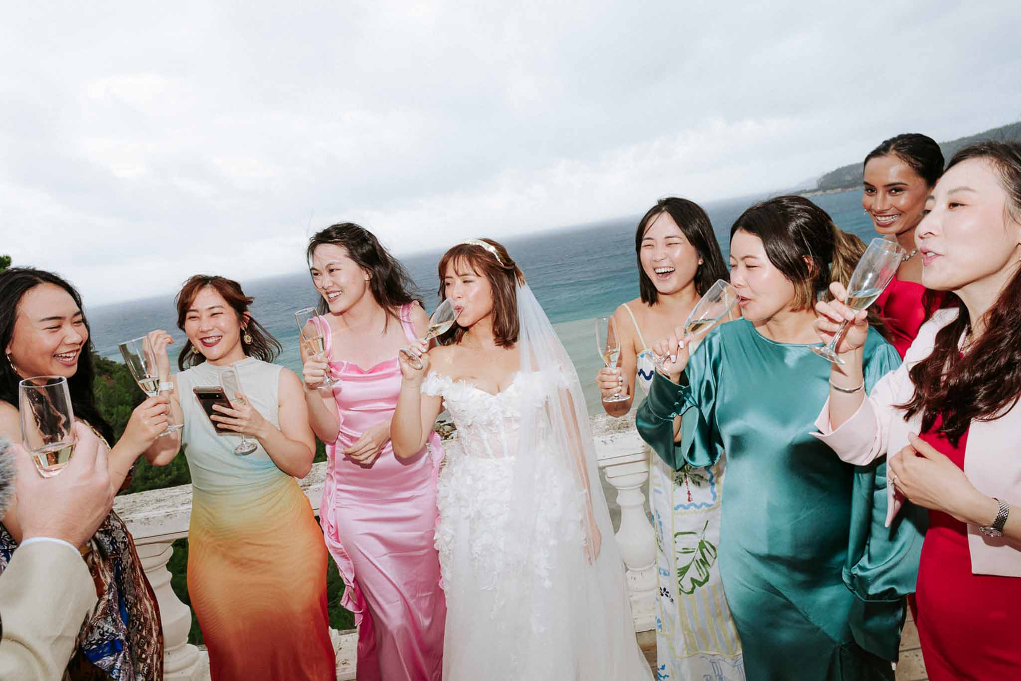 Bride and eight guests sharing champagne toast on sea-view terrace, colourful gowns against stone balustrade