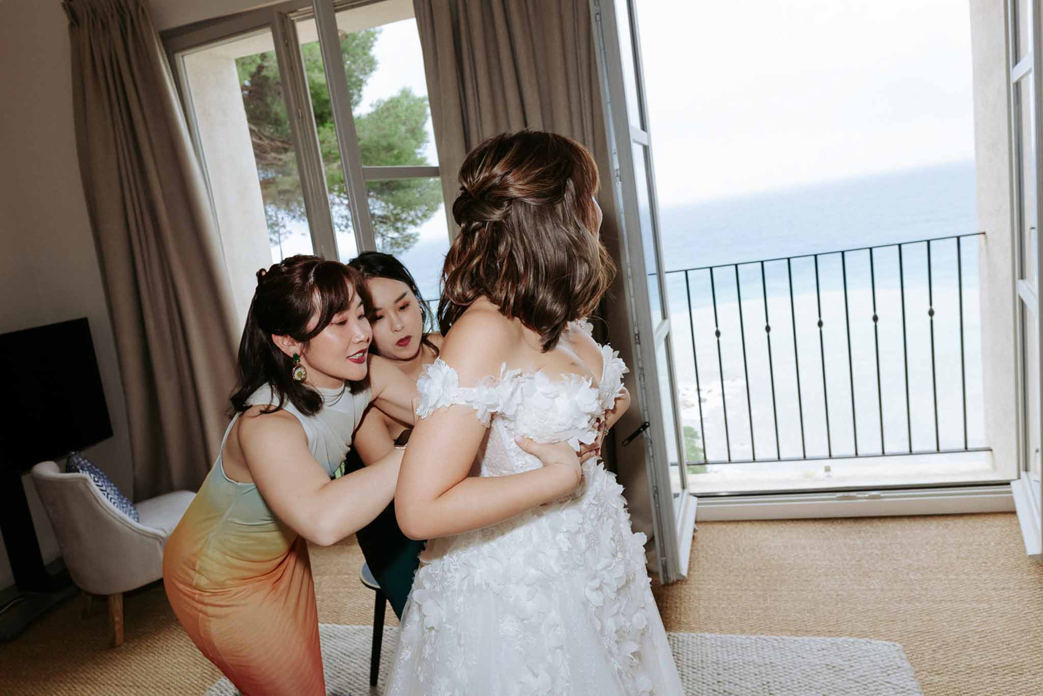 Two women helping bride into white floral-applique gown in room overlooking the sea through balcony doors