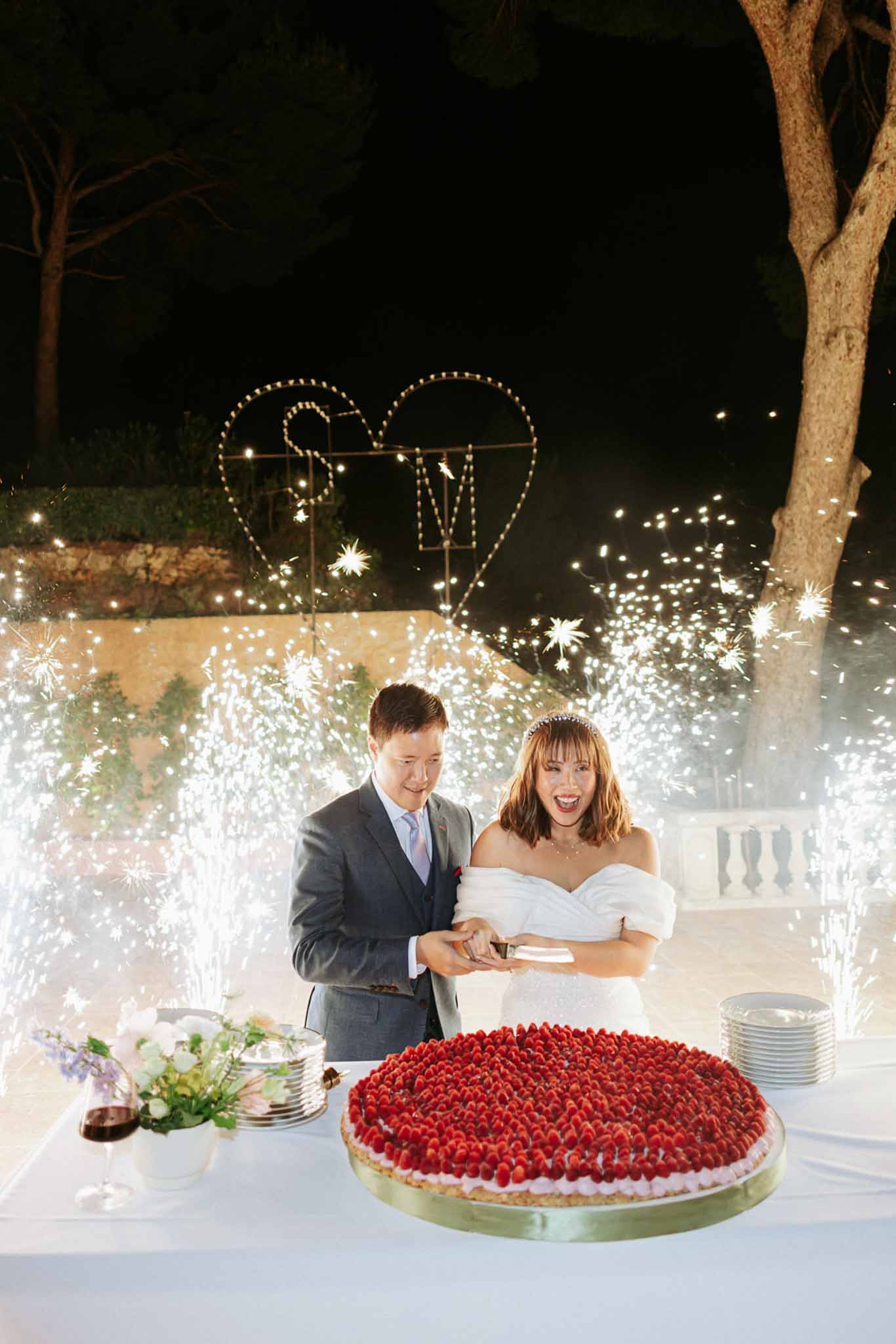Couple laughing while cutting strawberry cake at night with sparkler pyrotechnics and heart initials