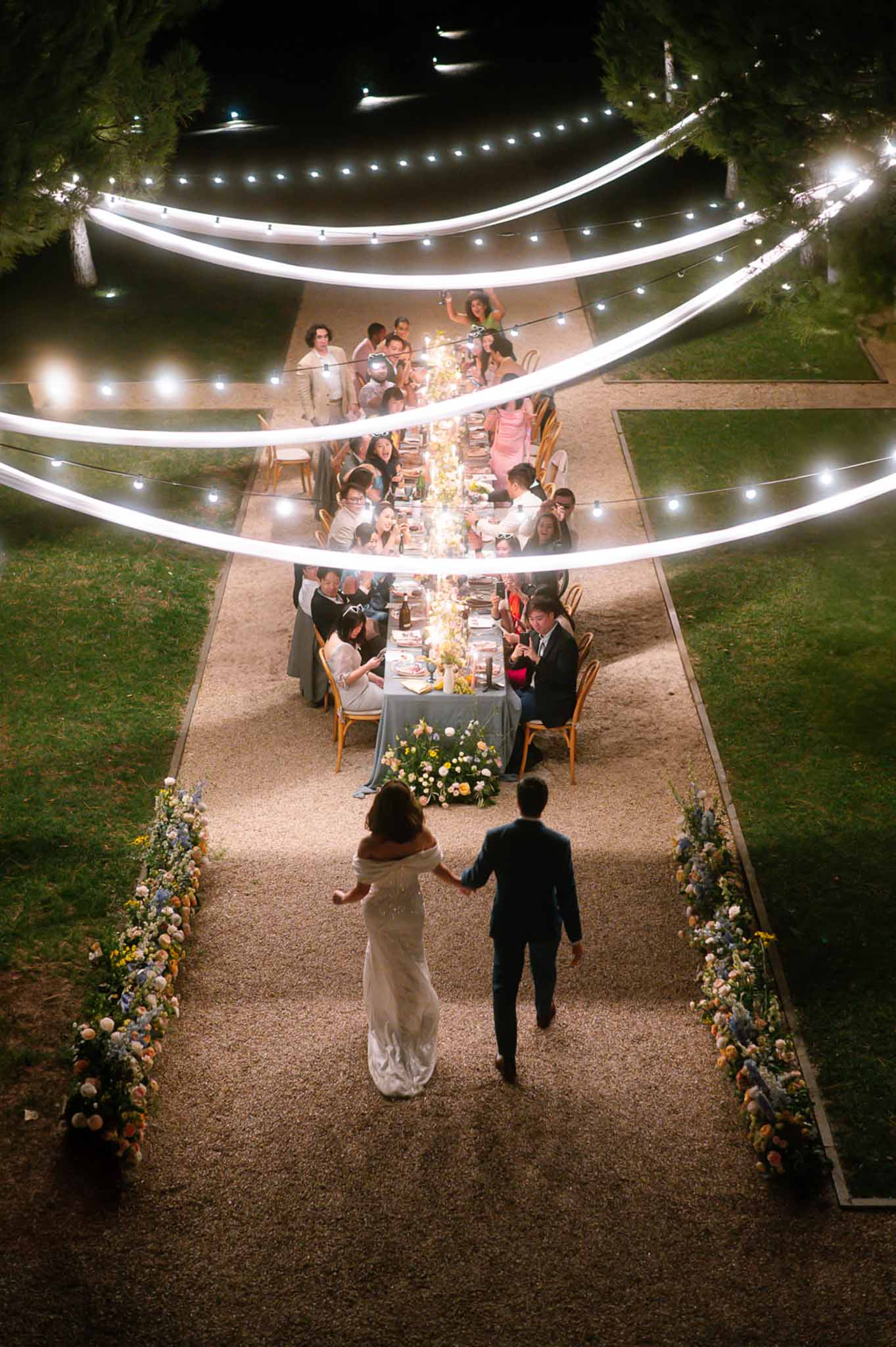 Night aerial of couple approaching candlelit blue table with yellow flowers under draped fairy light canopy