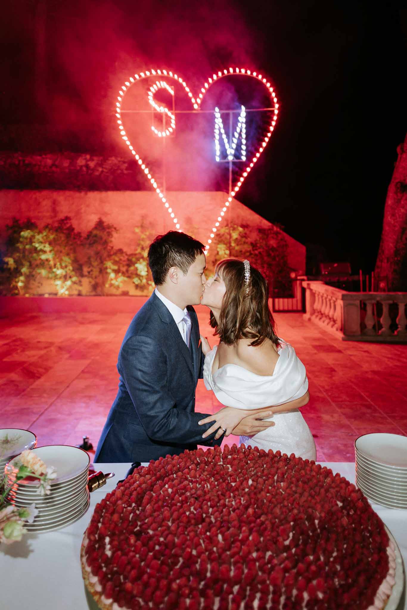 Couple kissing during cake cutting with raspberry tart and illuminated heart sign with red smoke