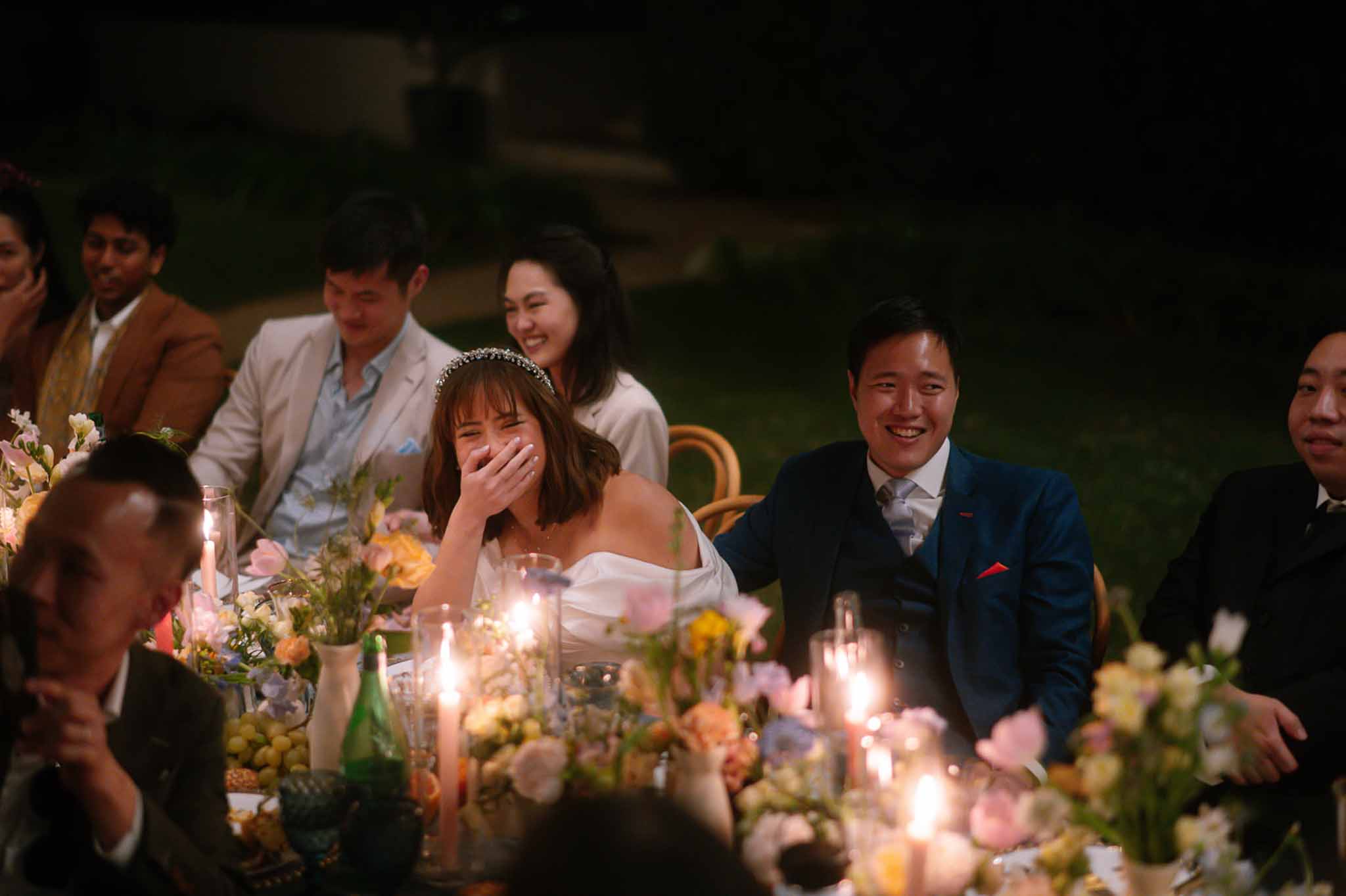Bride and groom laughing during reception speech at candlelit outdoor table with garden-style florals
