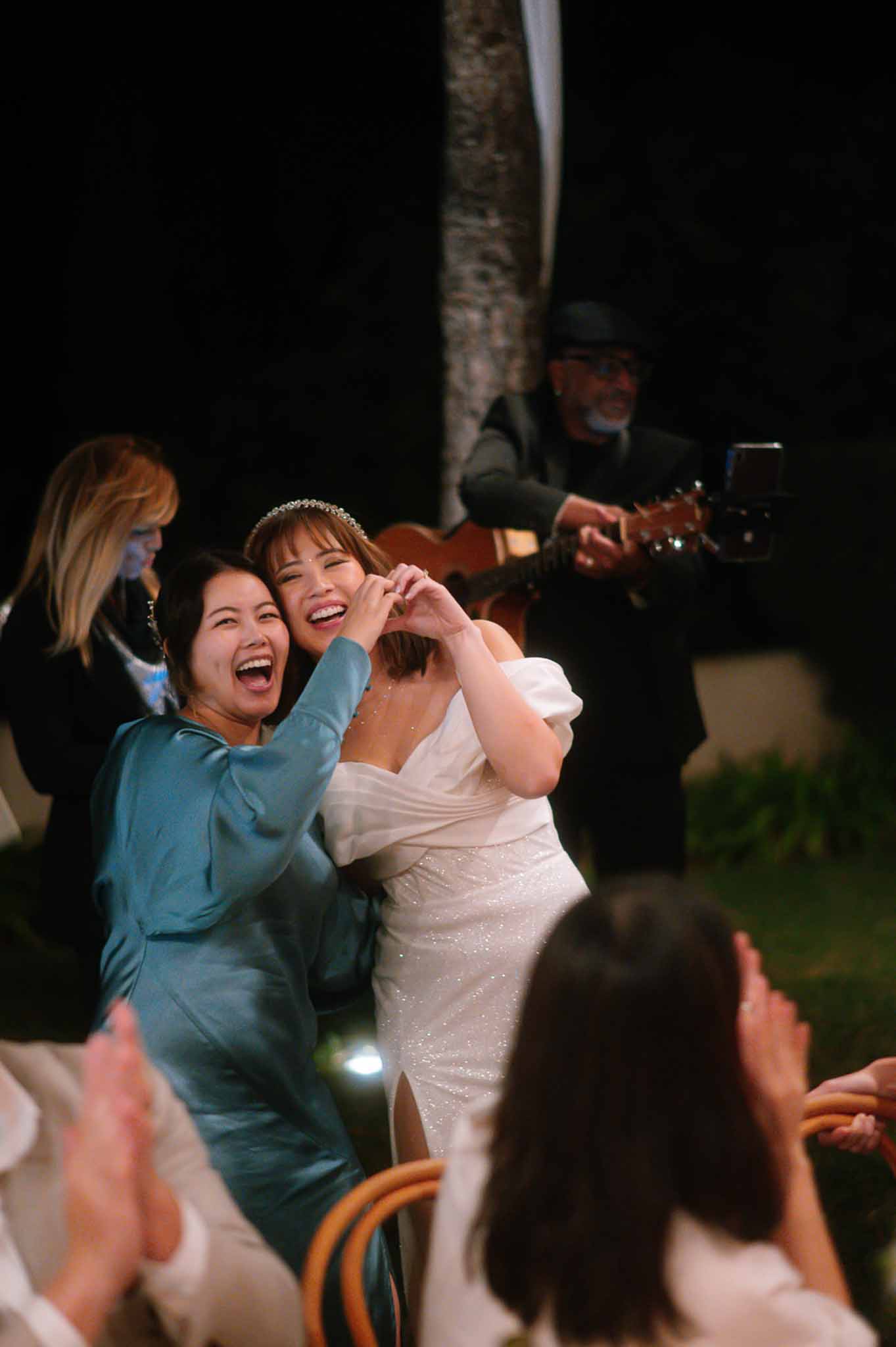 Bride in sparkle gown and tiara making heart hands with guest as guitarist performs at nighttime reception