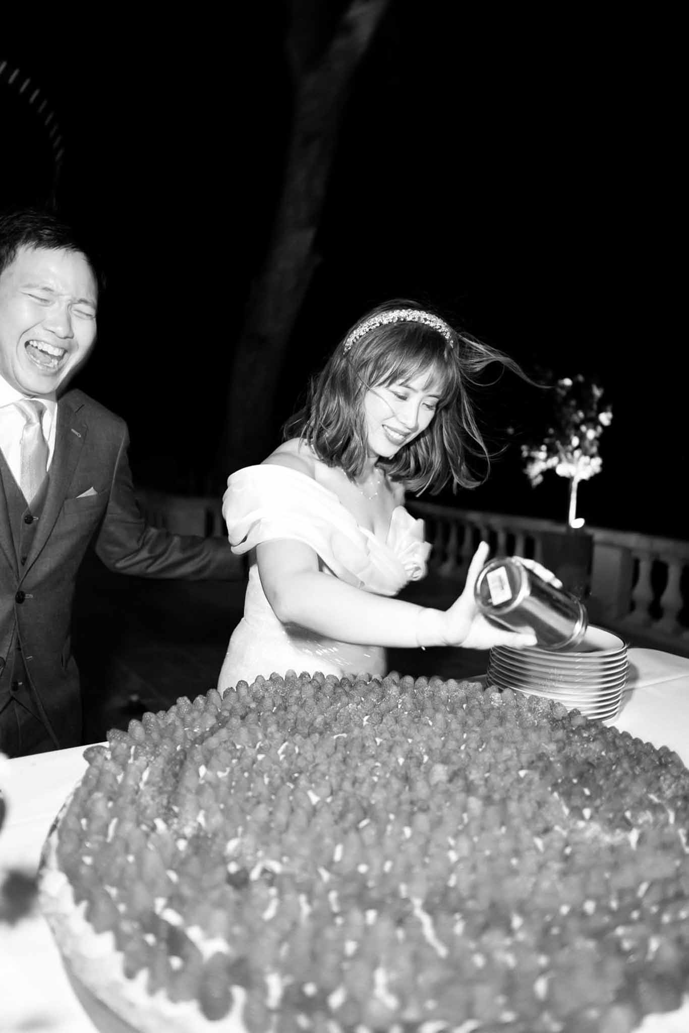 Bride pouring liquid onto strawberry-covered cake as groom laughs at outdoor evening reception in B&W