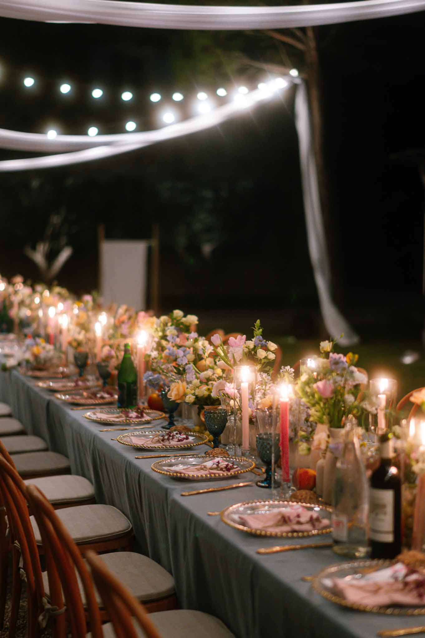 Evening banquet table with dusty blue linen, pastel tulip runner, taper candles, and globe string lights