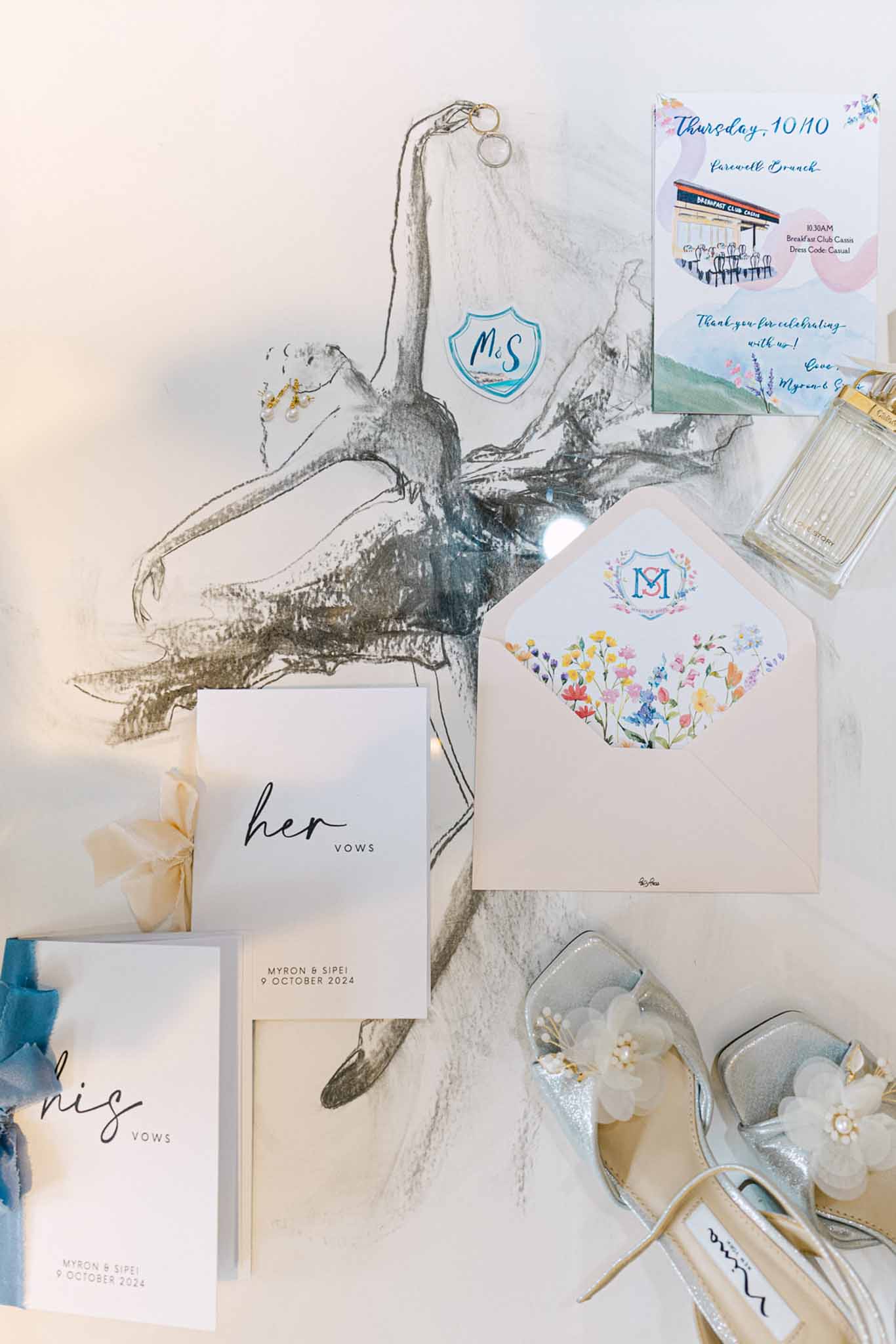 Flat lay of wedding stationery, vow booklets, silver glitter heels, pearl earrings, and blush envelope with monogram