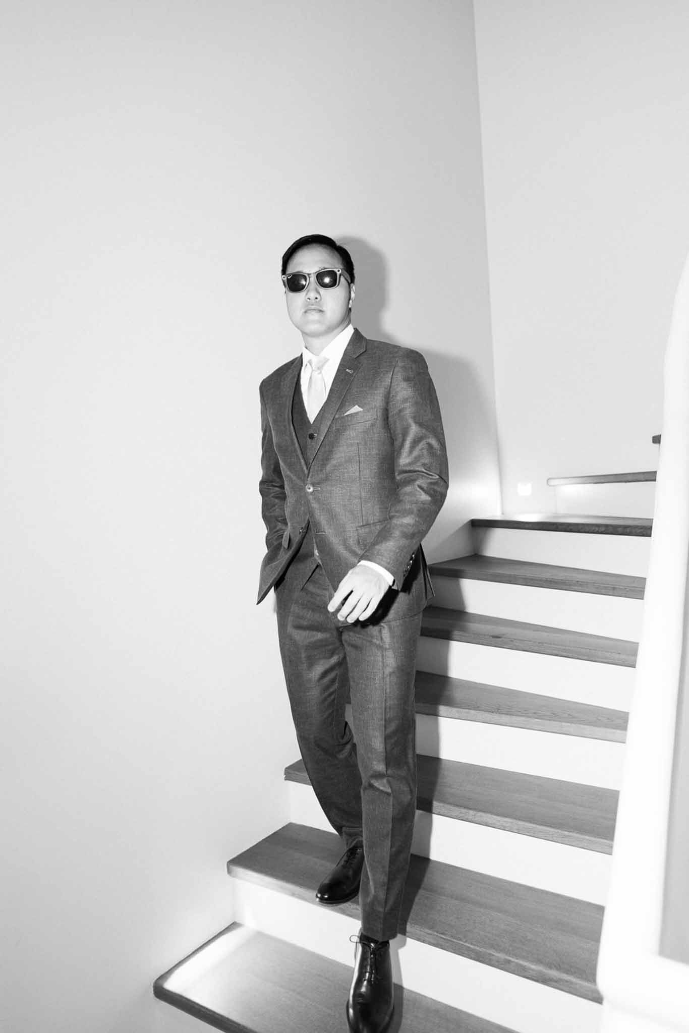 Black and white full-length portrait of a groom in a three-piece suit and sunglasses on a modern staircase