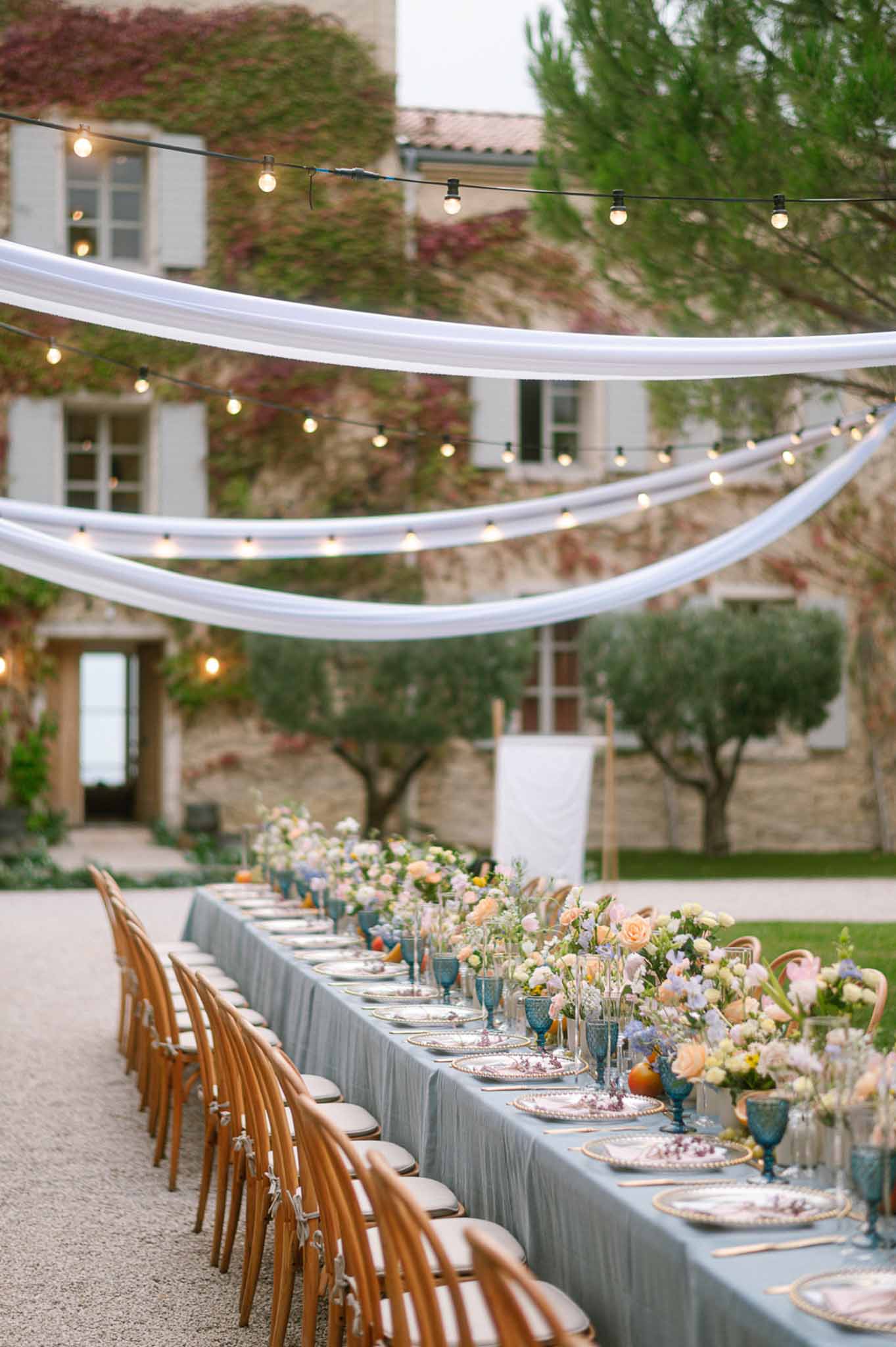 Long table with peach rose runner, teal goblets, and Edison lights on Provencal stone courtyard
