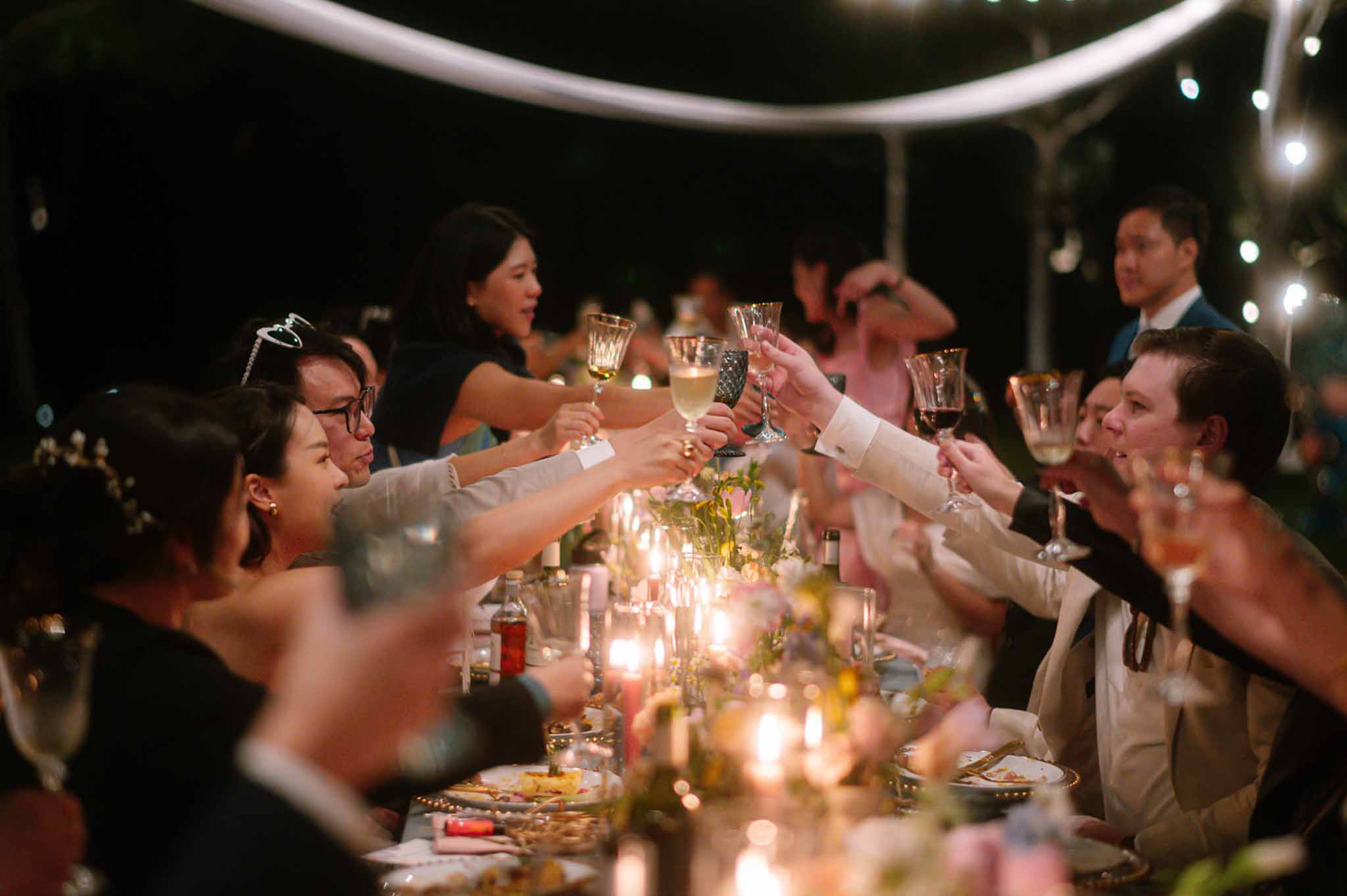 Guests raise glasses in candlelit toast at long reception table under fairy lights at night