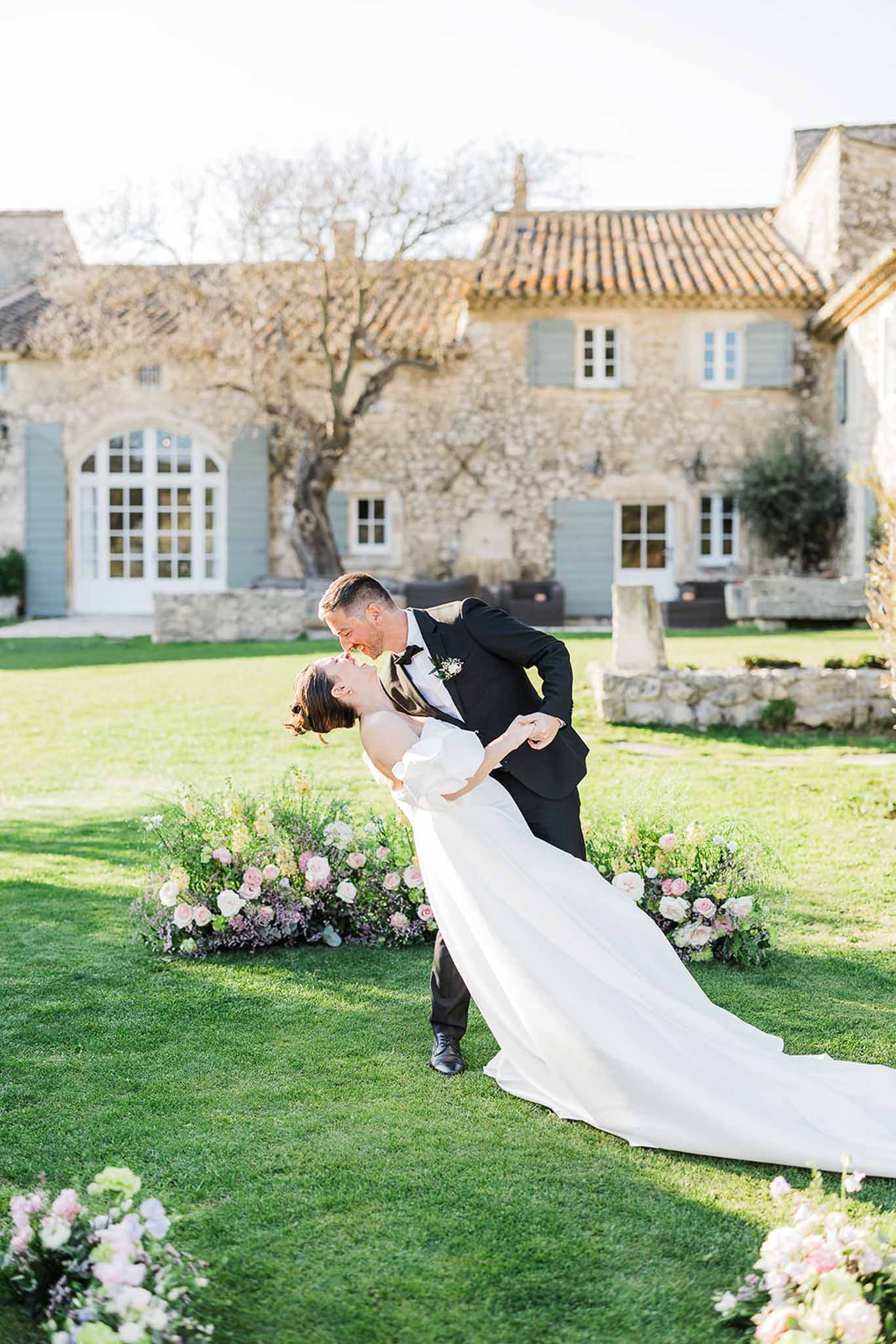Groom dips bride for romantic kiss on lawn of Provençal stone manor with hydrangea arrangements