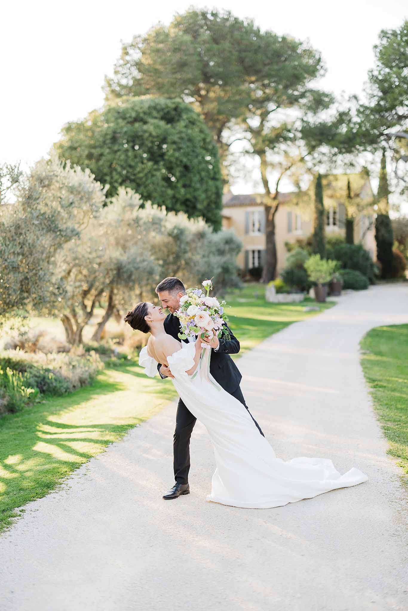 Groom lifting bride on tree-lined gravel pathway at European-style estate wedding venue