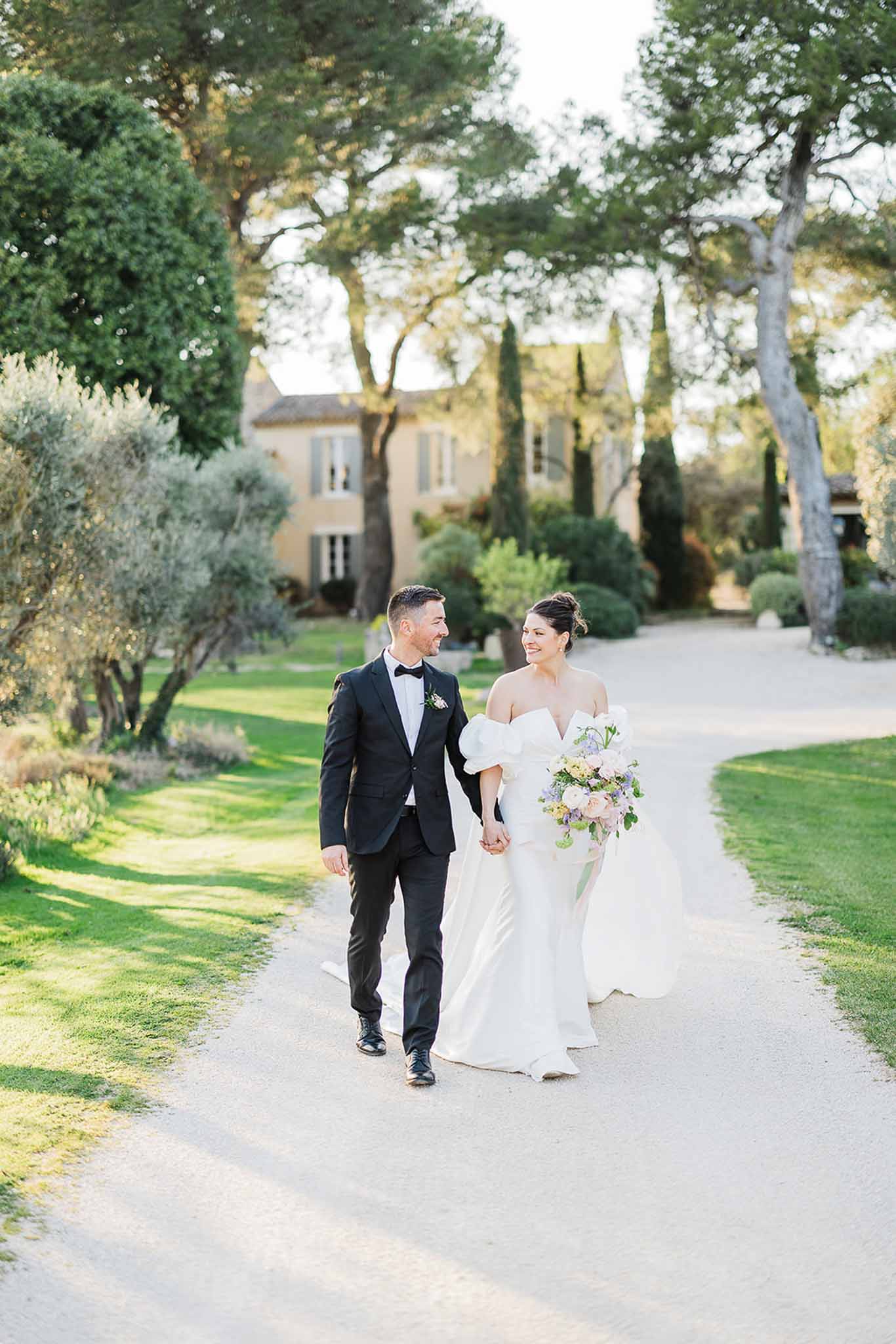 Bride and groom walking hand-in-hand down tree-lined driveway at European estate manor